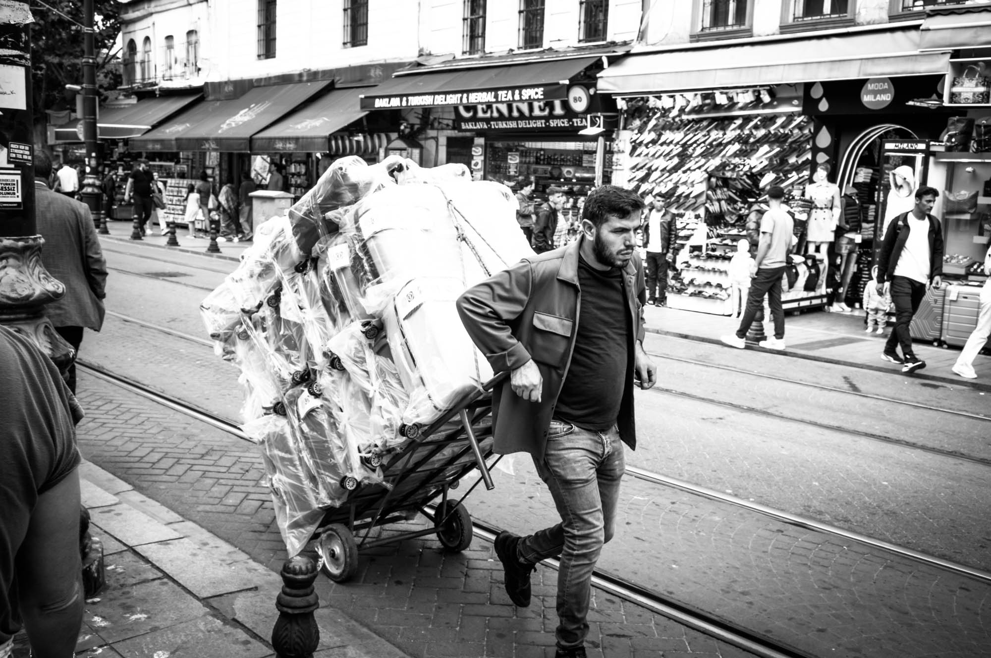 Man pulling a cart full of packages on a bustling street lined with shops. Black and white photo.