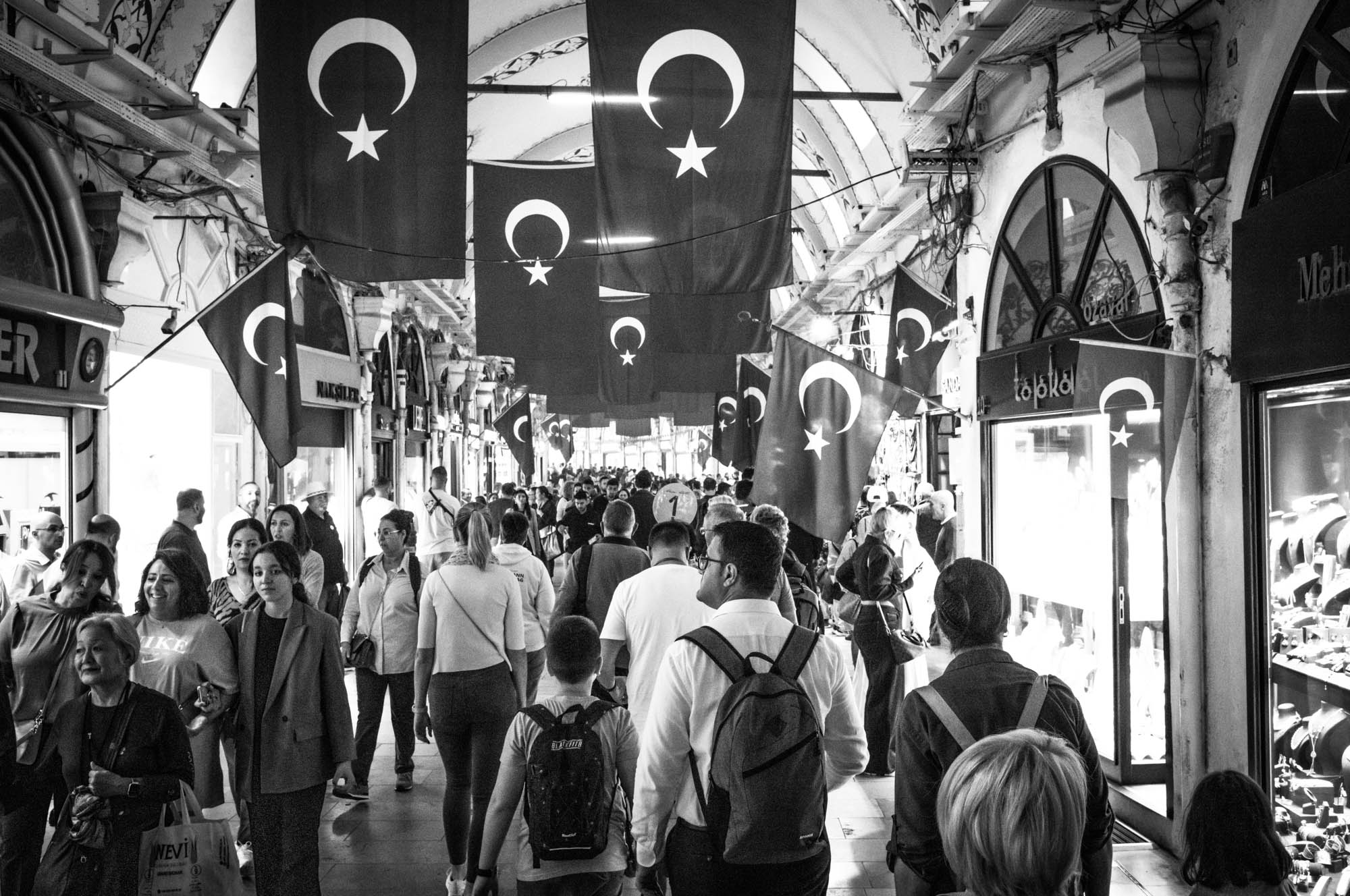 Busy market street in Istanbul's Grand Bazaar, filled with people and Turkish flags. Black and white photograph.