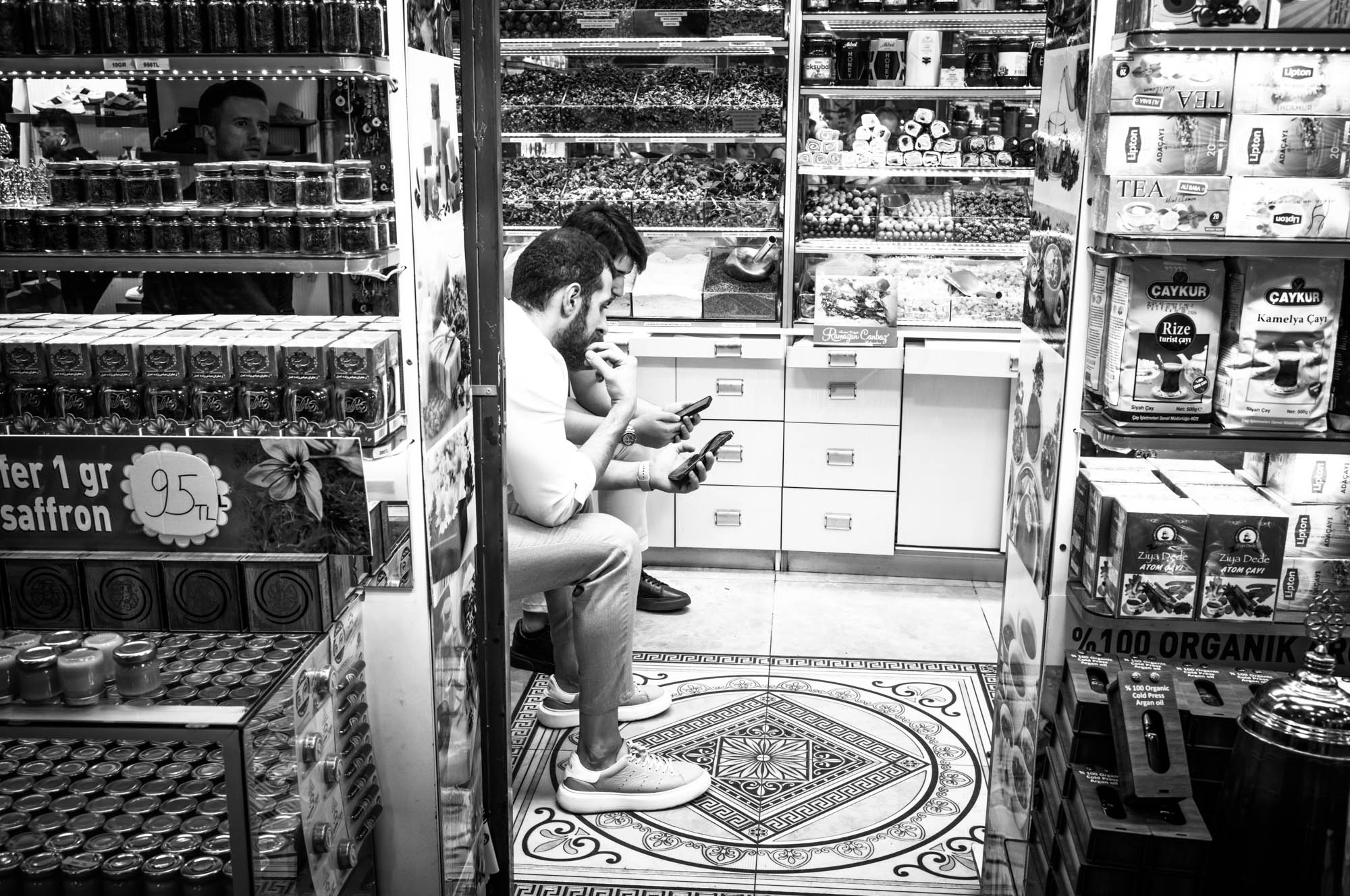 People in spice shop browsing phones, surrounded by jars and packages, black and white.