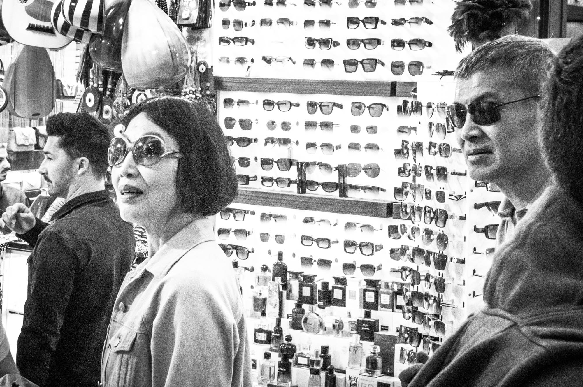 Shoppers browse sunglasses in a store filled with eyewear displays, captured in black and white.