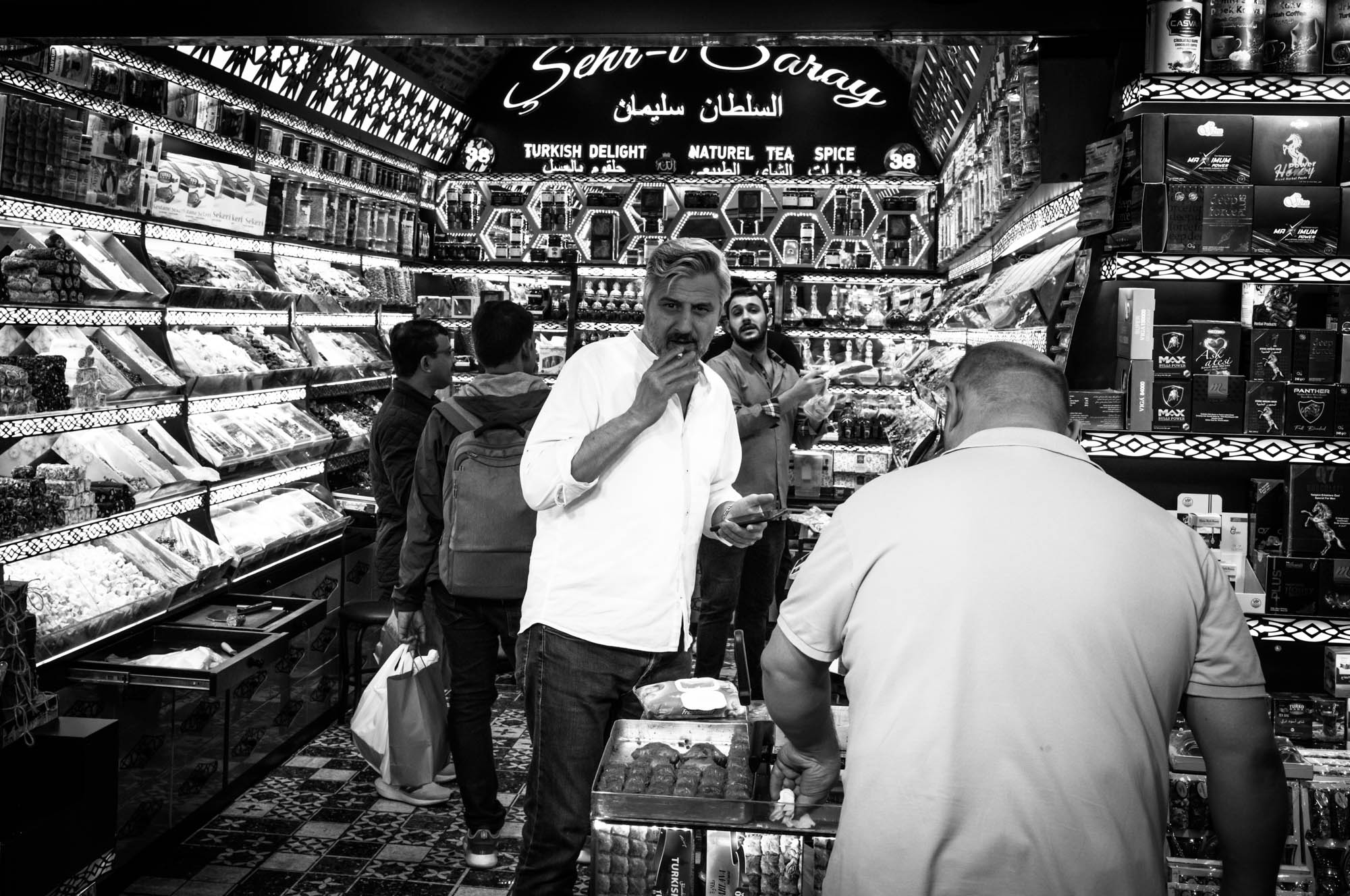 Man tasting sweets in a bustling Turkish delight shop, surrounded by colorful candies and vibrant decor.