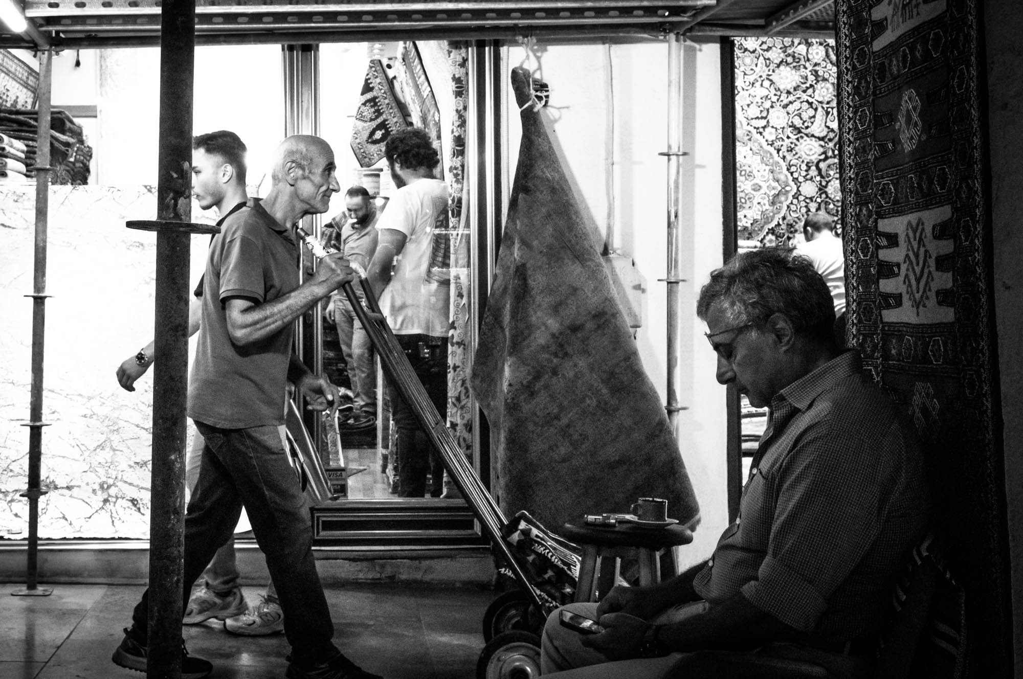 Men working and relaxing in a busy carpet shop, with intricate patterns in the background, black and white photo.