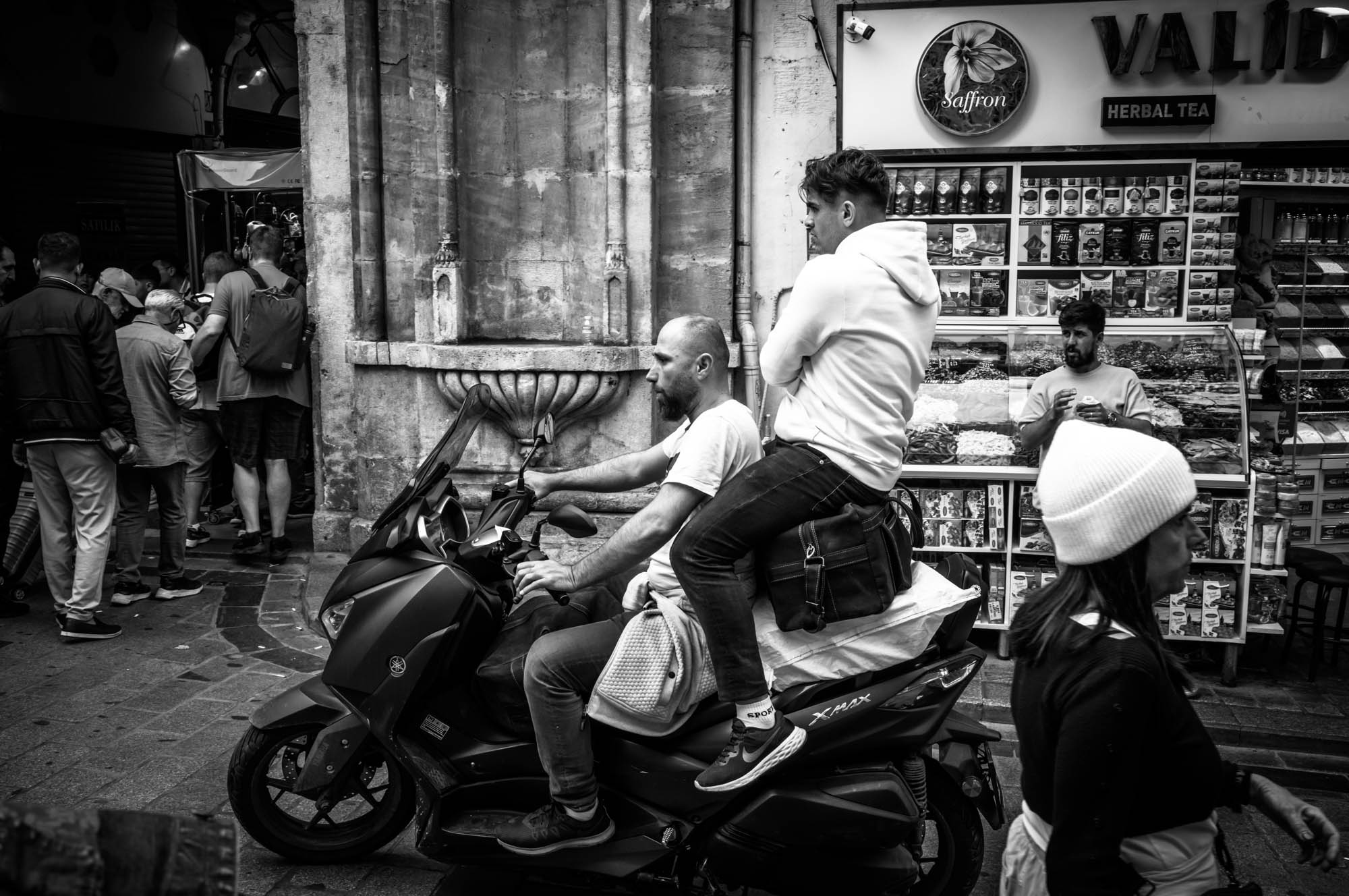Two men on a scooter in a bustling street, with a tea shop and pedestrians in the background. Black and white street scene.