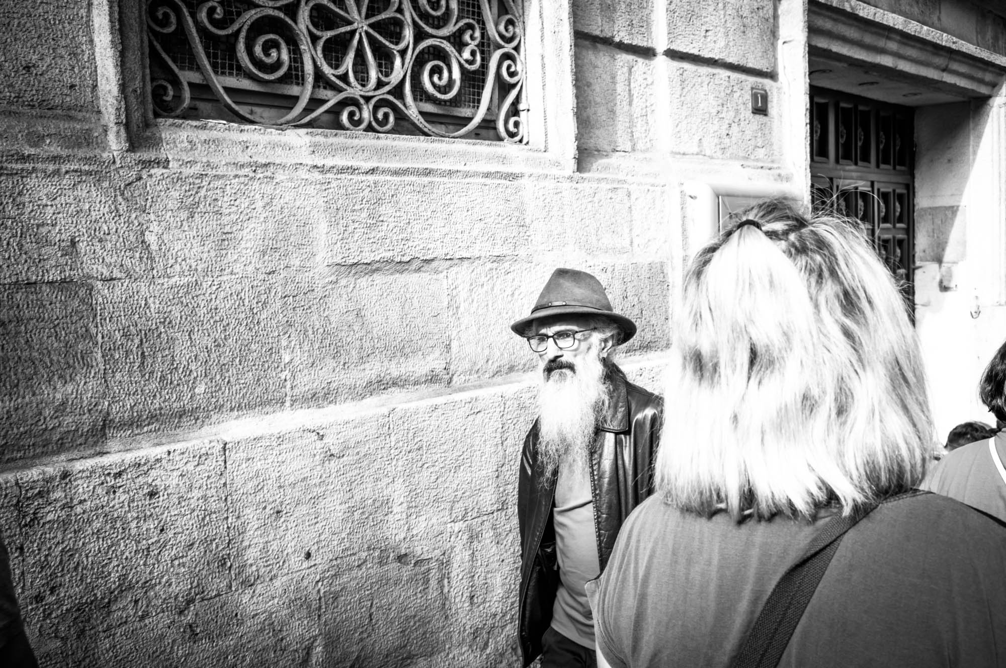 Man with beard and hat standing by ornate wall, black and white street scene.
