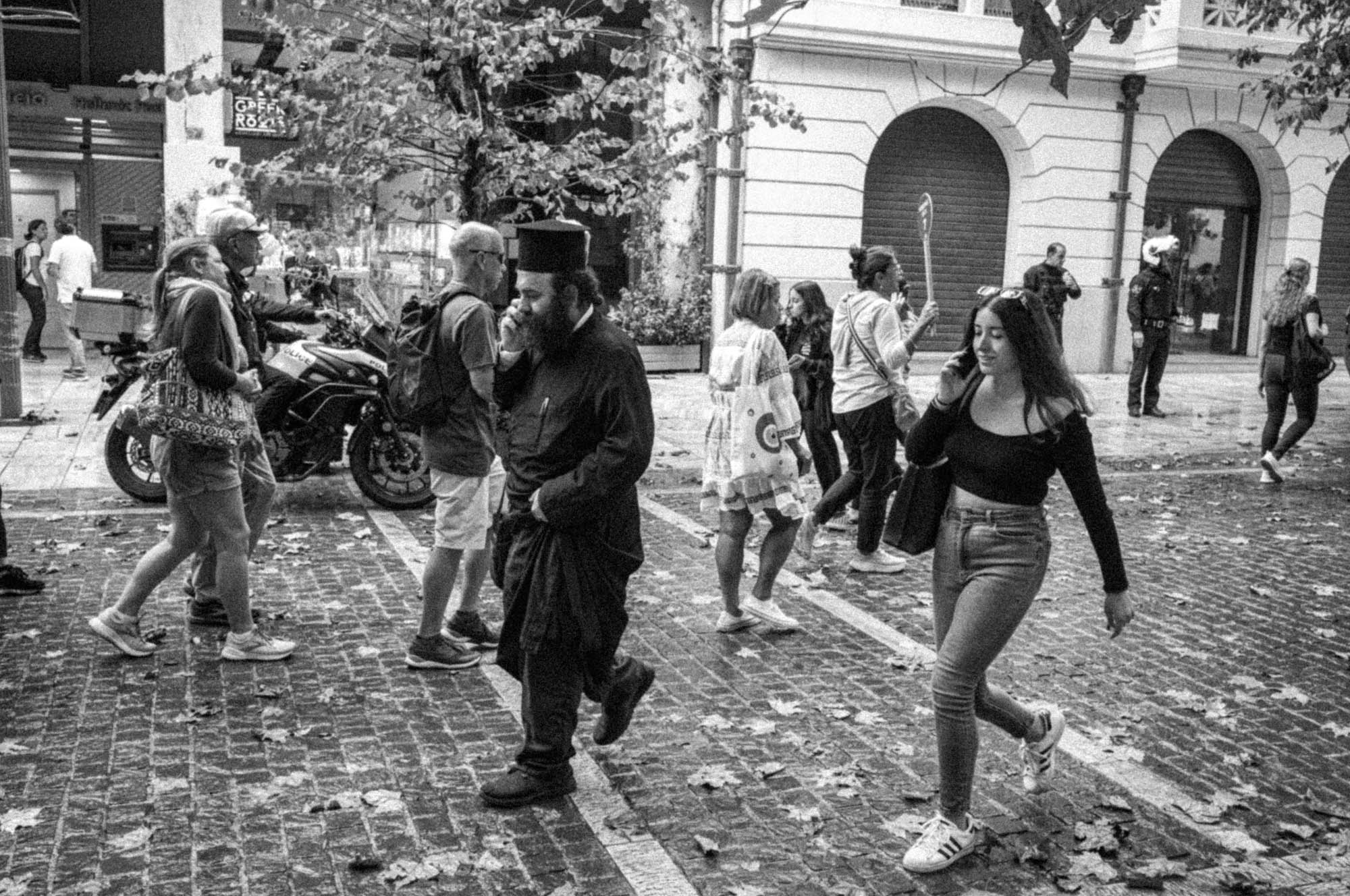 Black and white street scene with pedestrians walking, some using phones, and a motorcycle in the background.