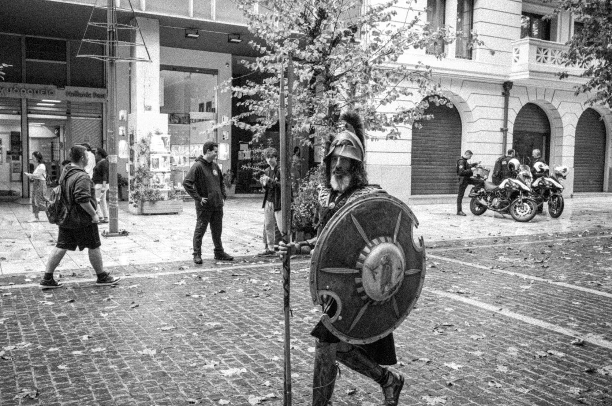 Historical reenactor in armor with shield on a city street; people and motorcycles in the background.