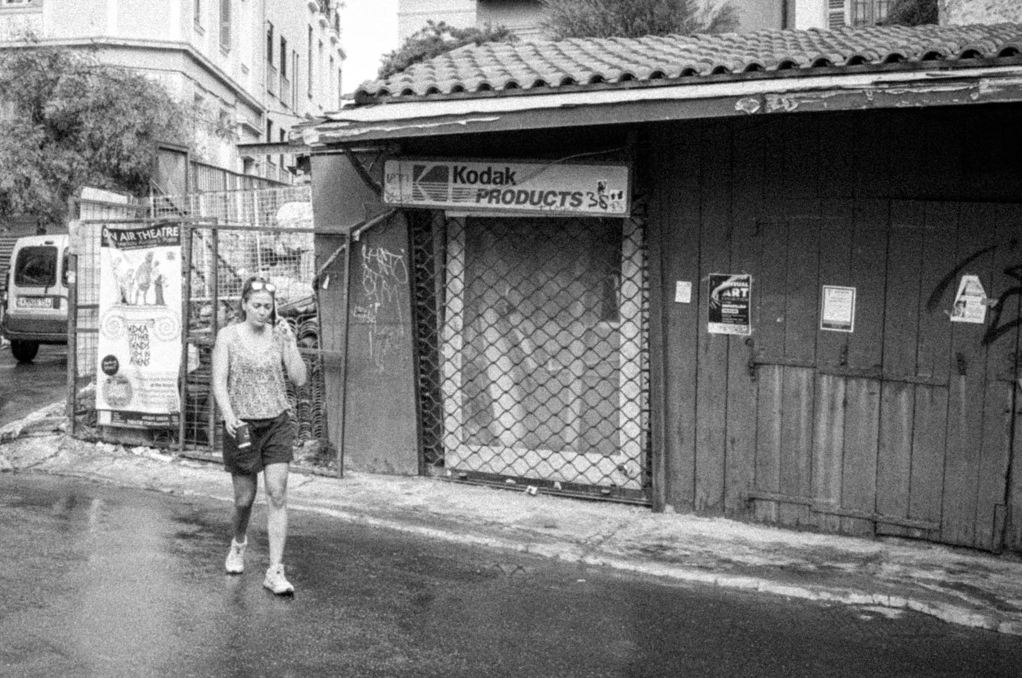 Person walking past an old Kodak store with a closed gate, urban street in black and white.