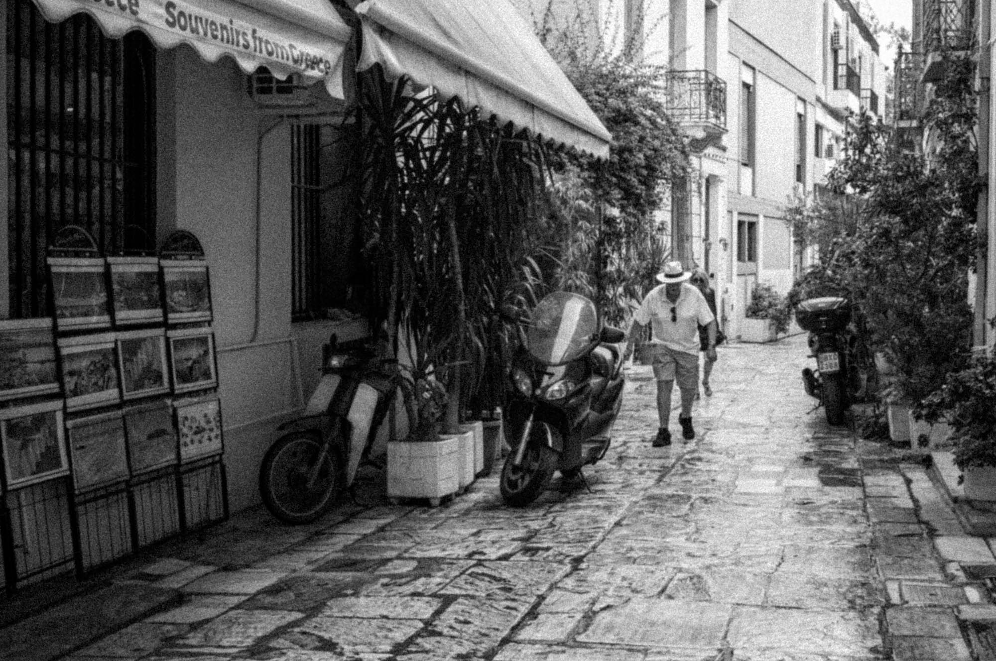 Black and white photo of a narrow Greek street with souvenir shop and scooters.