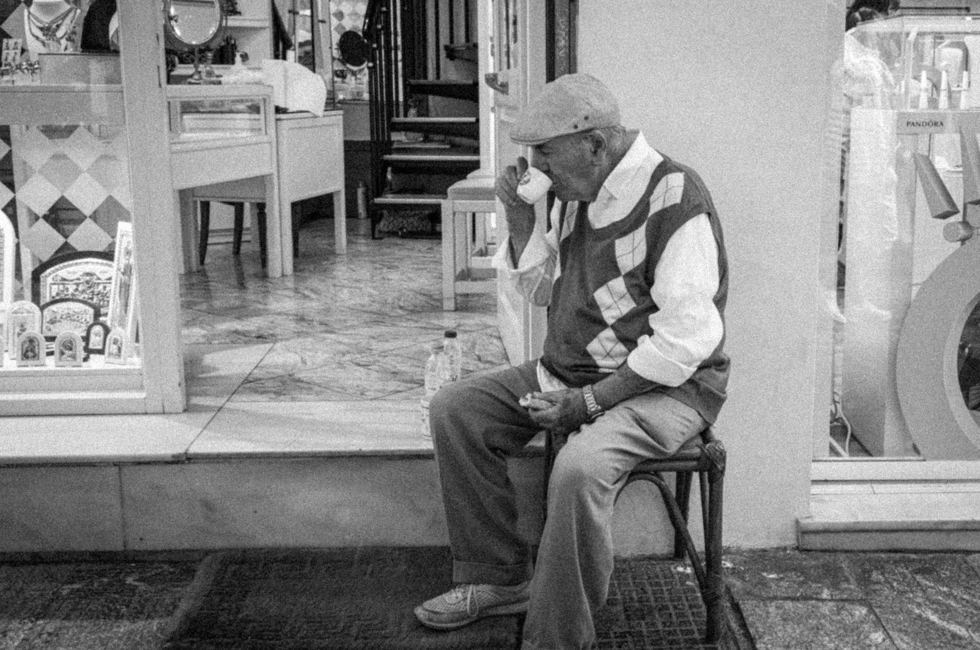 Elderly man in hat enjoys espresso outside a shop, sitting on a stool, captured in black and white photography.
