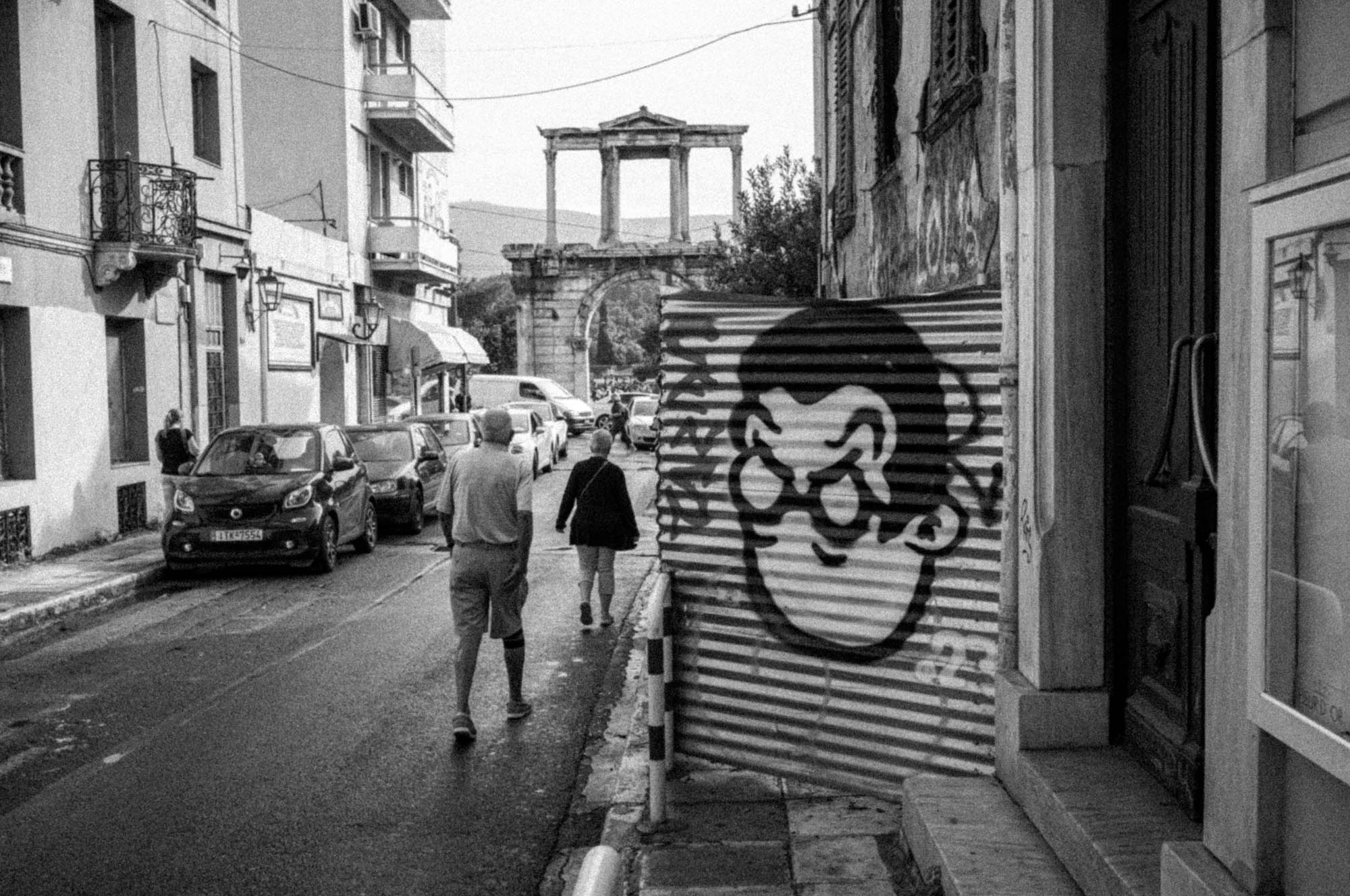 Street with graffiti art, people walking, ancient archway in background, black and white urban landscape.
