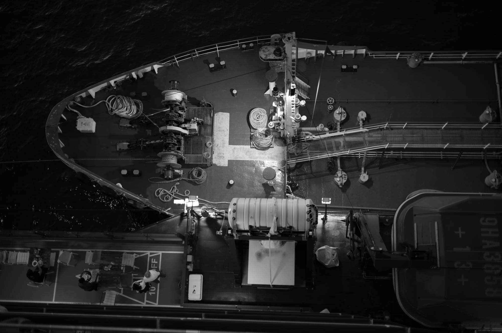 Aerial view of a ship's deck at night with ropes and equipment, illuminated against the dark sea background.