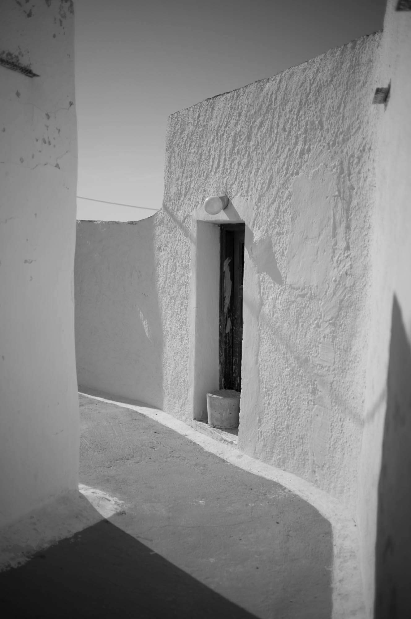 Narrow alley with whitewashed walls and a small doorway in bright sunlight, conveying minimalistic architecture.