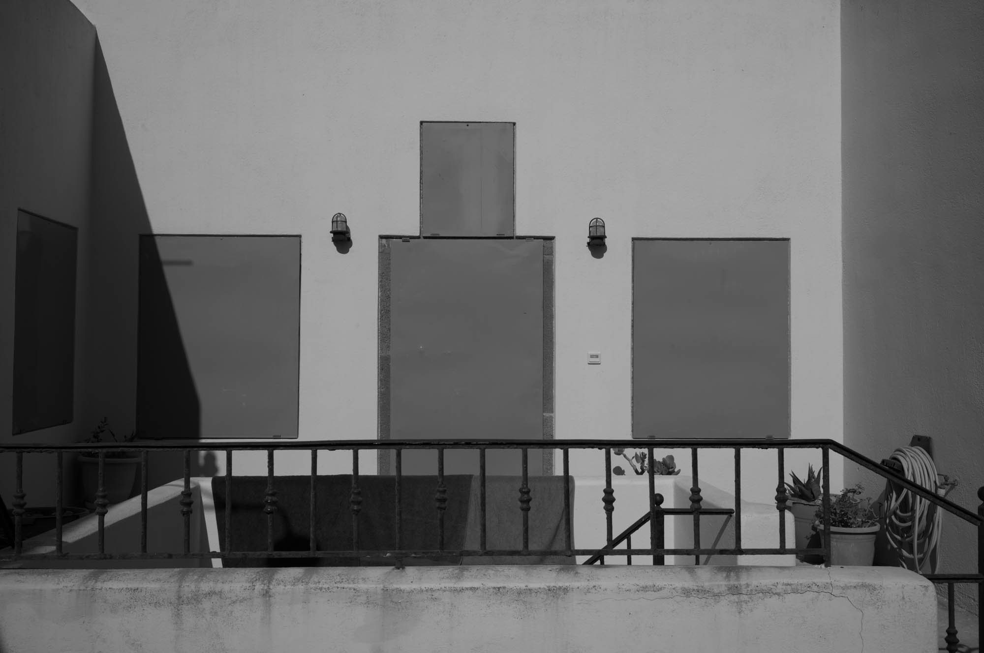 Minimalistic building facade with boarded-up windows, metal railing, and shadow patterns in black and white.
