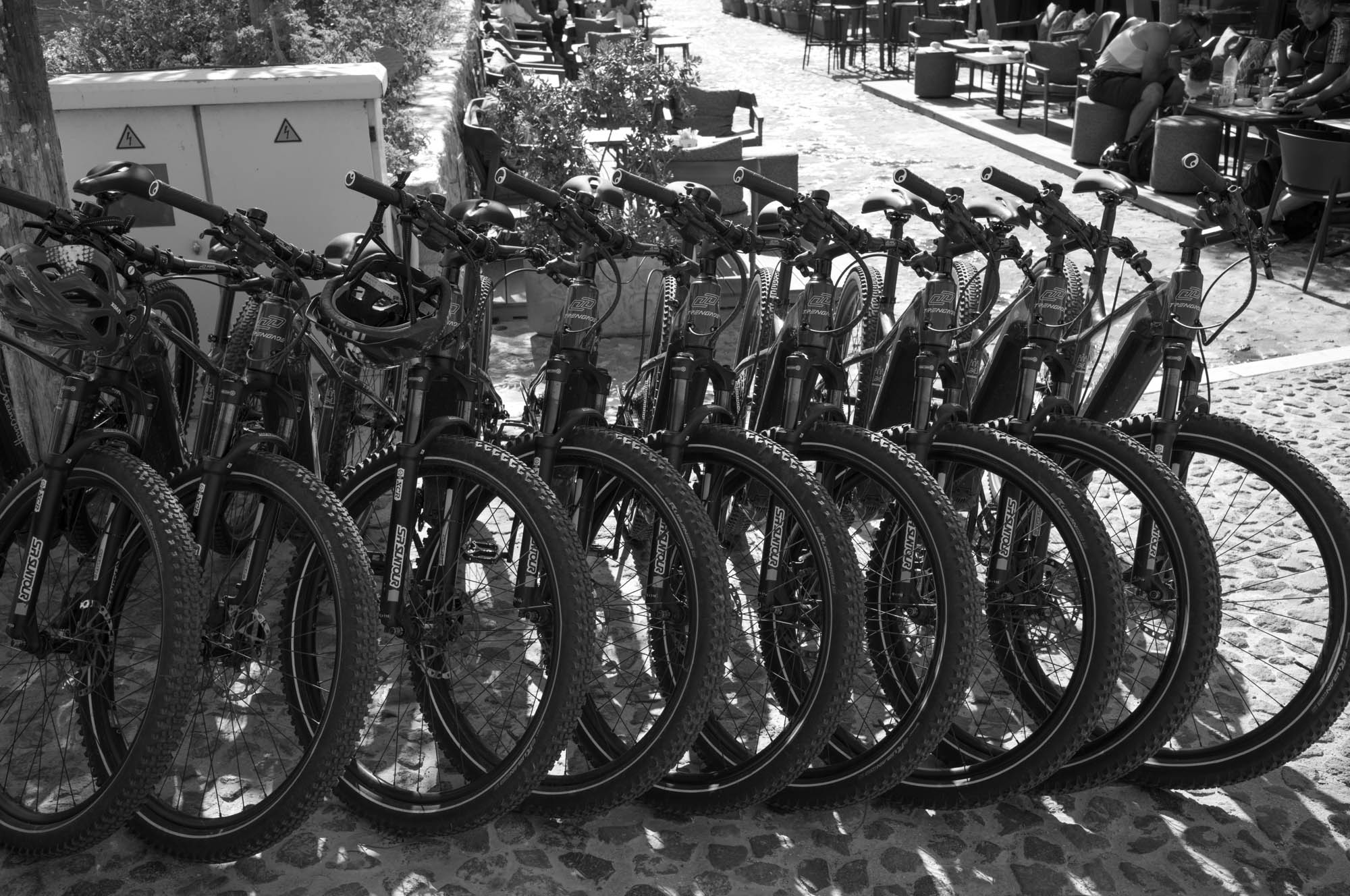 Row of mountain bikes parked on a cobblestone path near an outdoor seating area. Black and white photography.