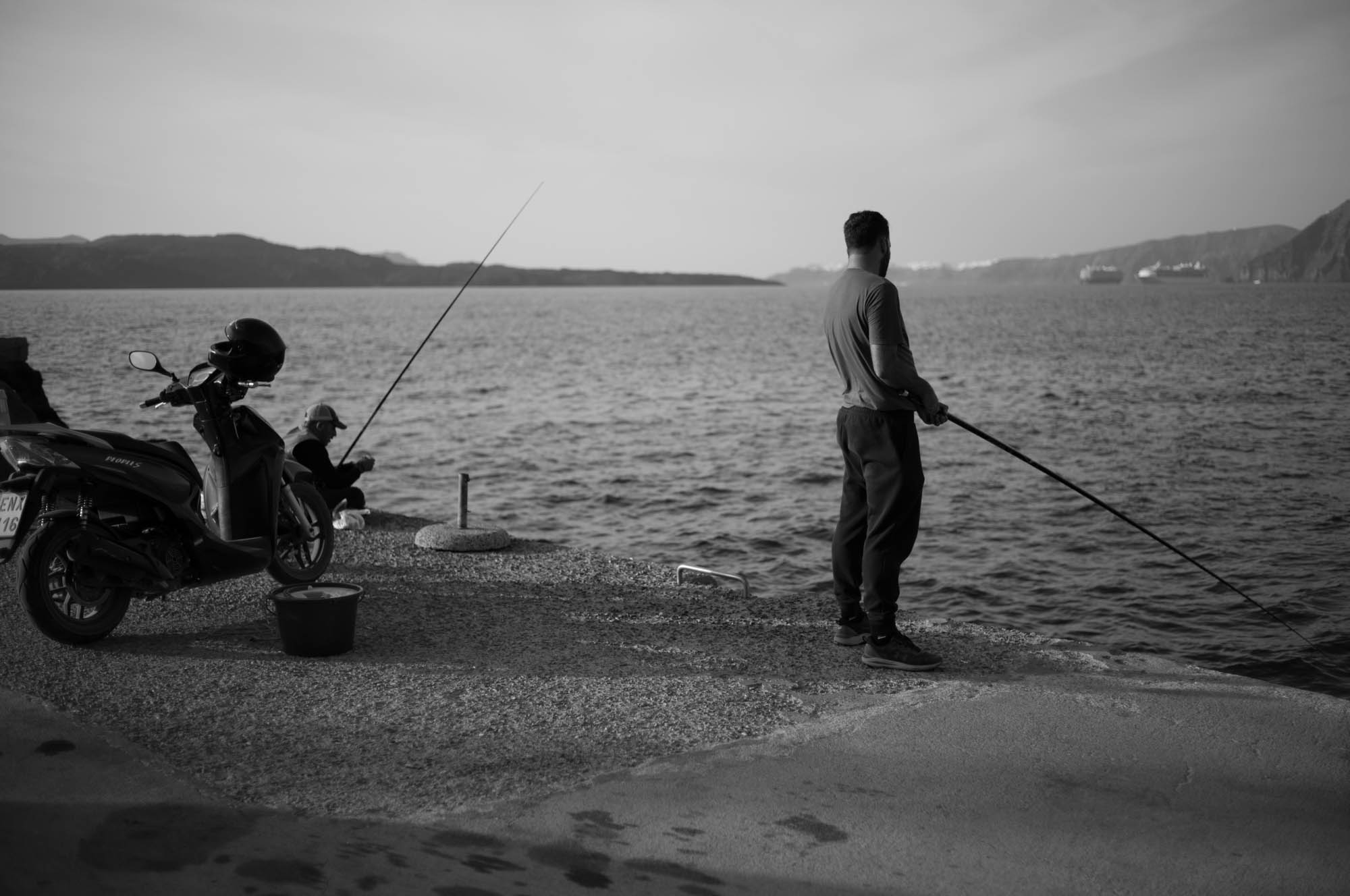 Two people fishing on a pier, with a motorcycle parked nearby, overlooking a calm sea.