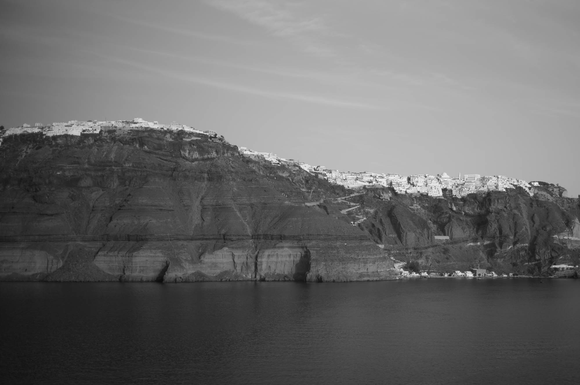 Black and white photo of a coastal cliff with white buildings perched on top, Santorini, Greece.