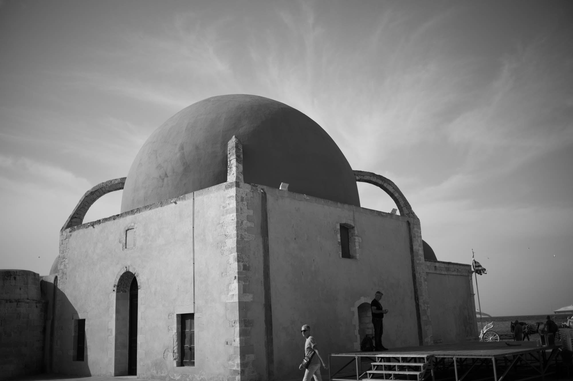 Monochrome photo of an ancient domed building with arches, set against a cloudy sky, people in the foreground.