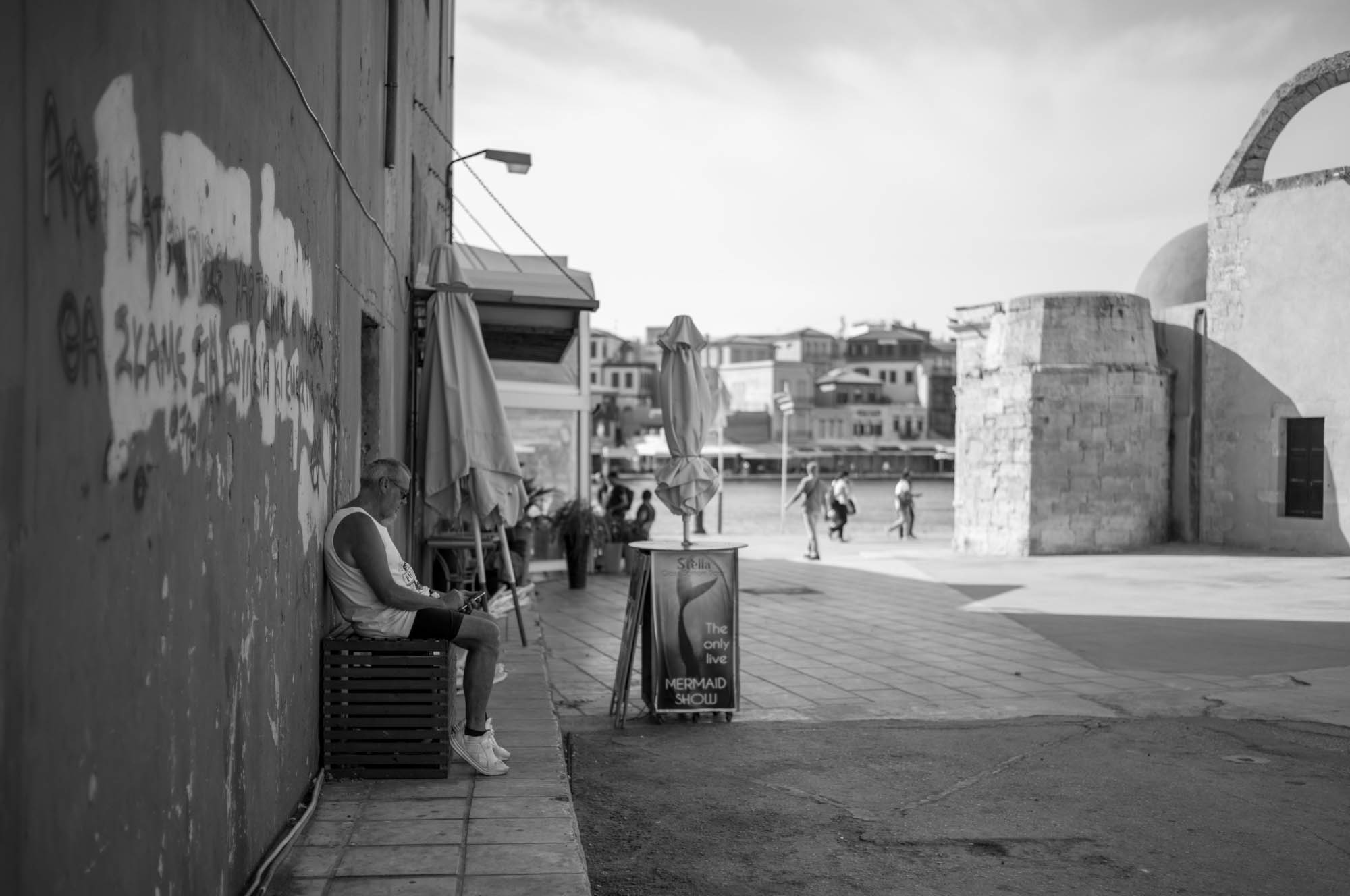 Black and white street scene by the waterfront, person sitting alone on bench near old building.