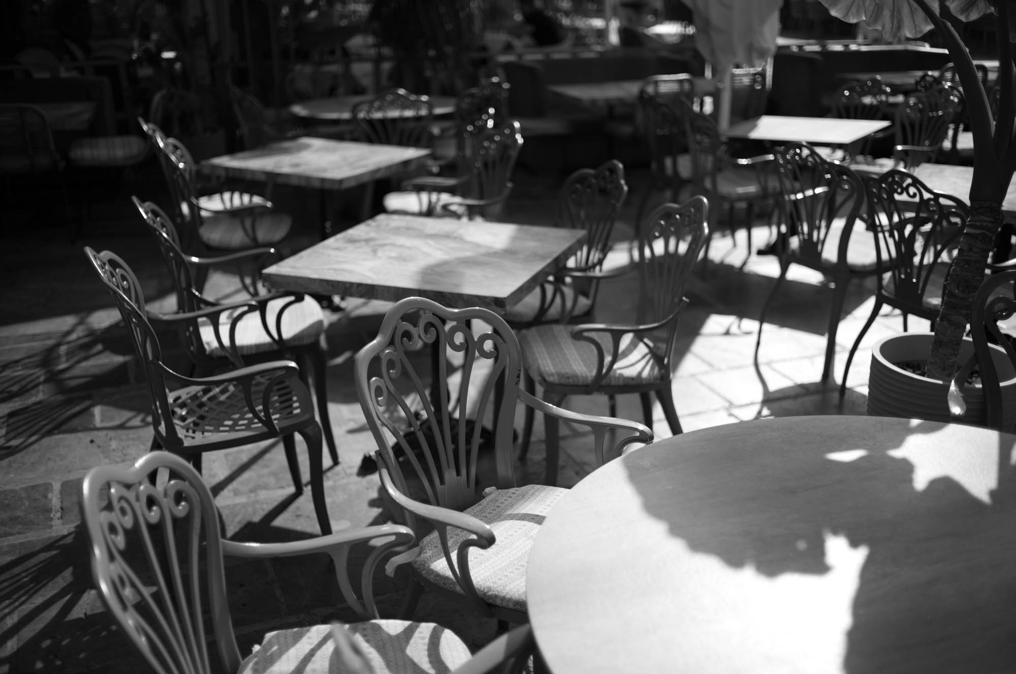 Black and white photo of empty outdoor cafe tables and chairs in the sunlight.