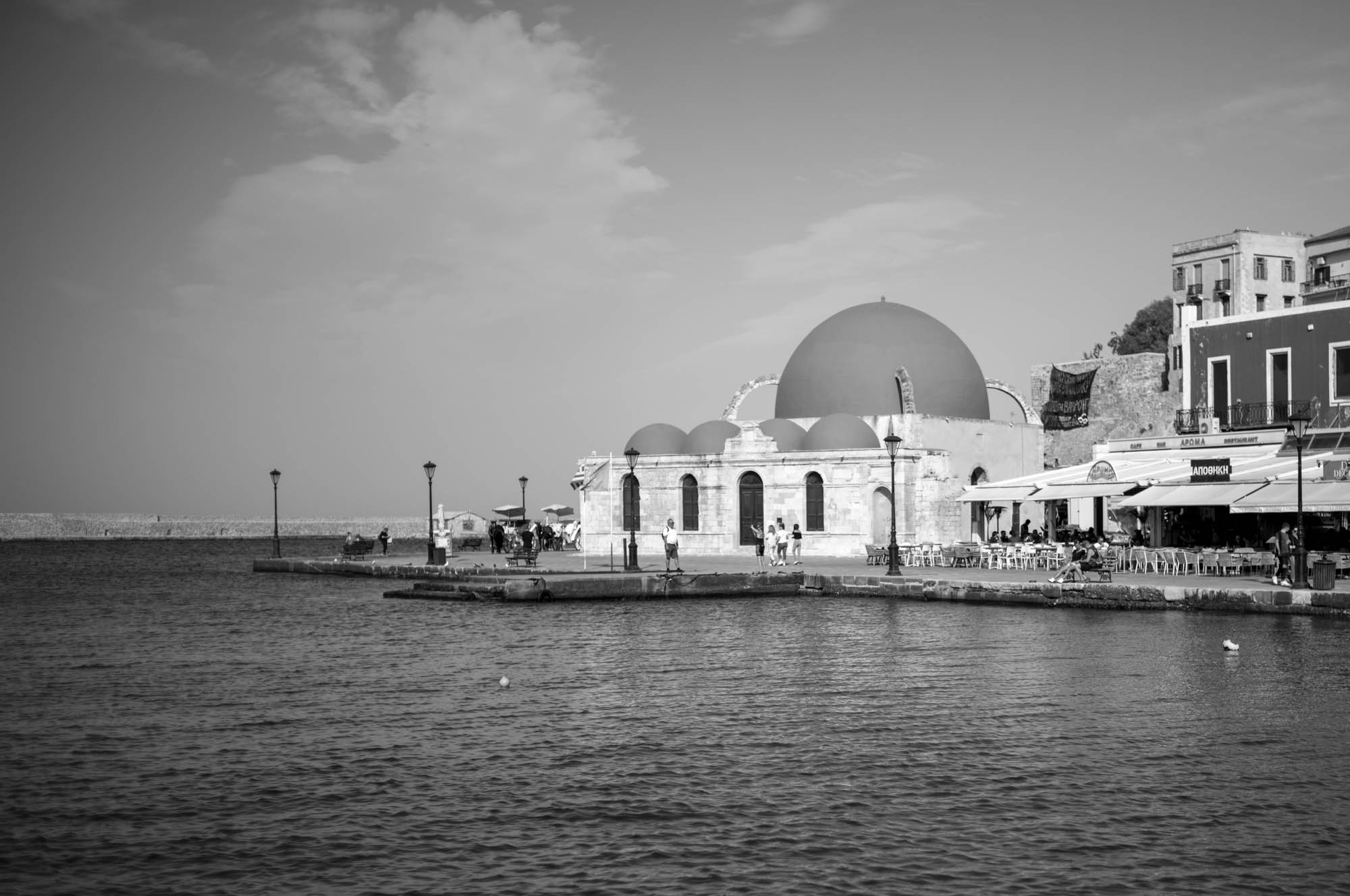 Historic building by the waterfront in black and white, featuring domes and a harbor setting with people strolling nearby.