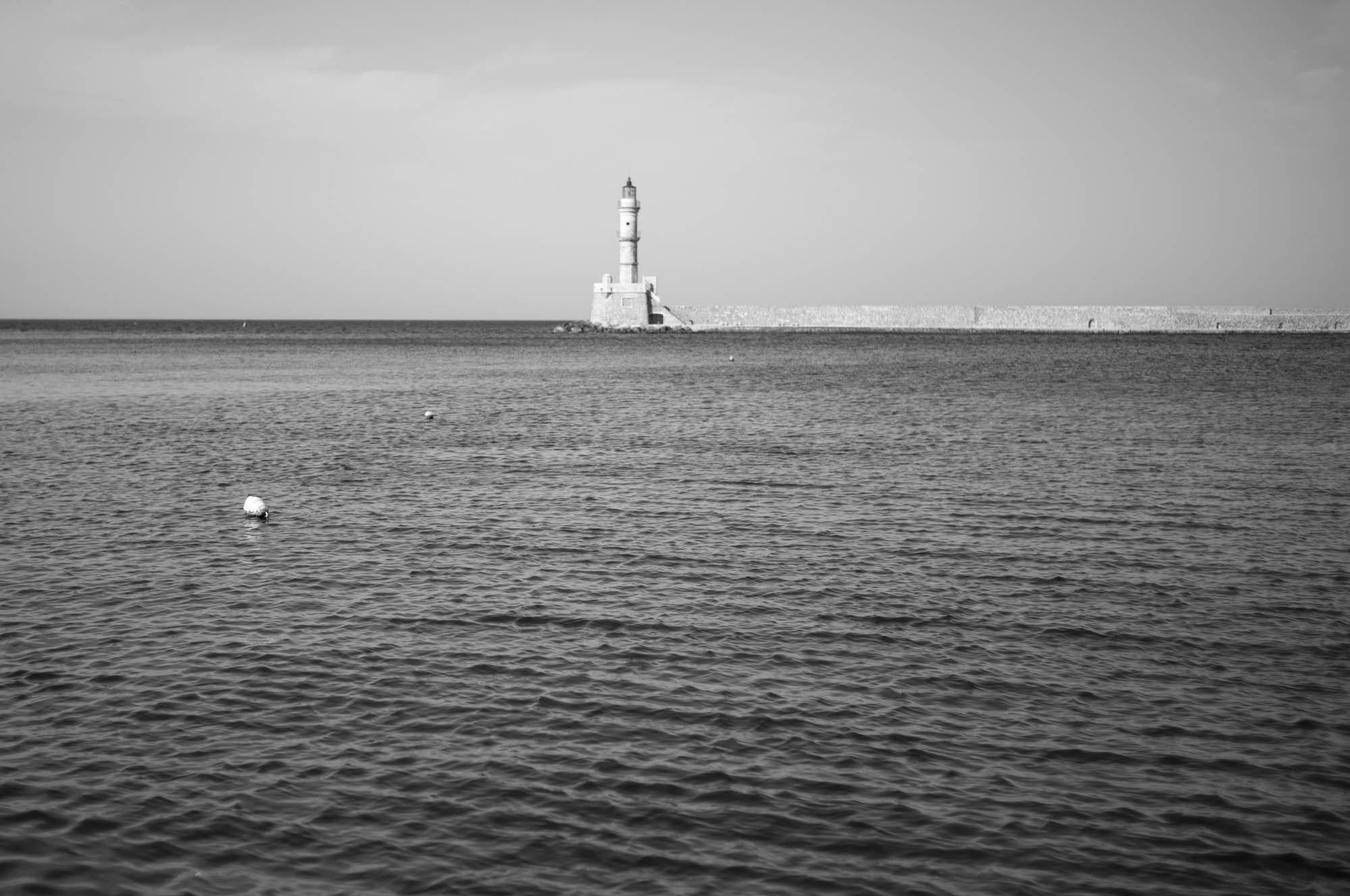 Black and white photo of a distant lighthouse on a calm sea, under a hazy sky.