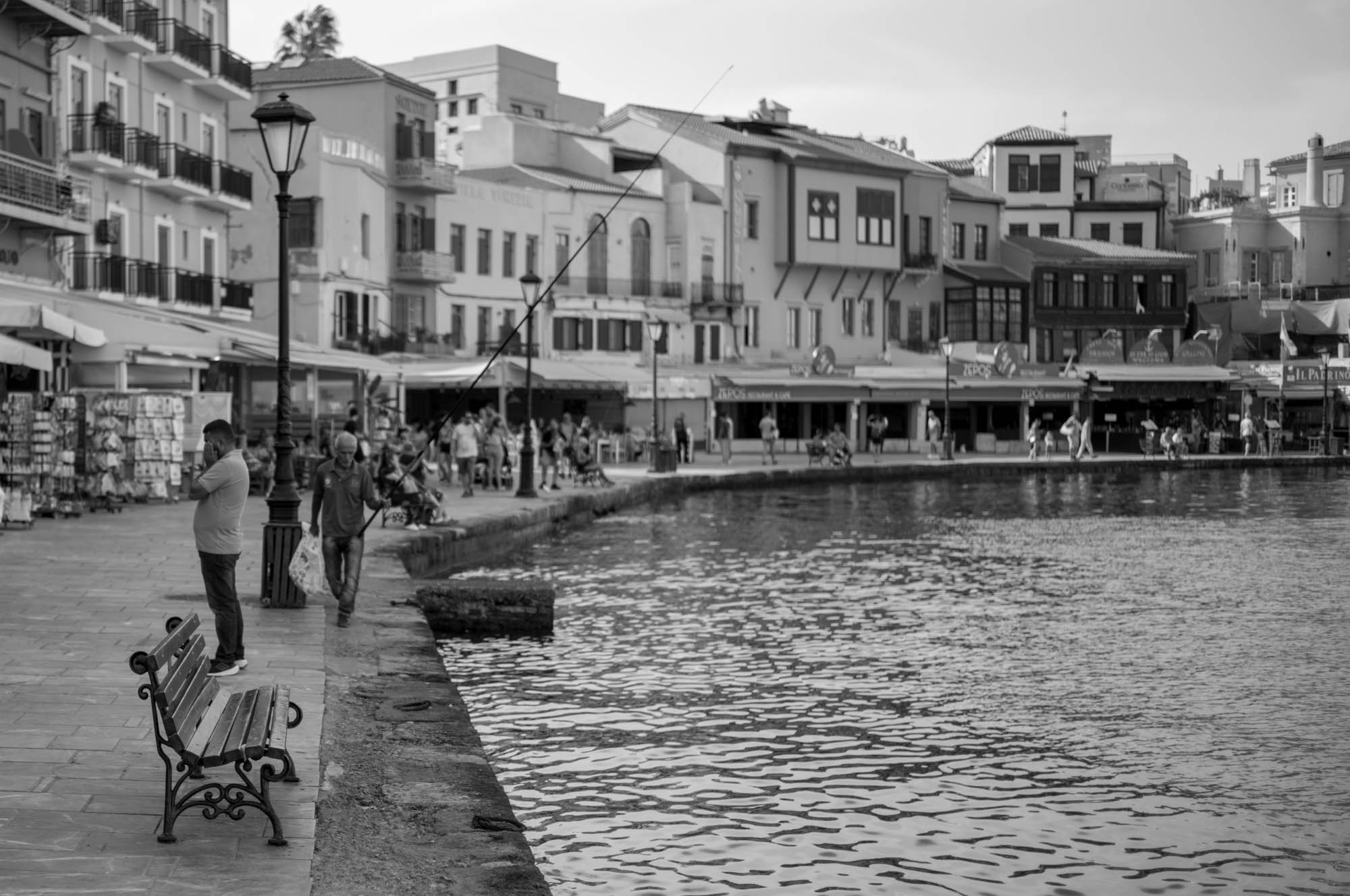 Black and white waterfront scene with people fishing and strolling along a promenade lined with shops.