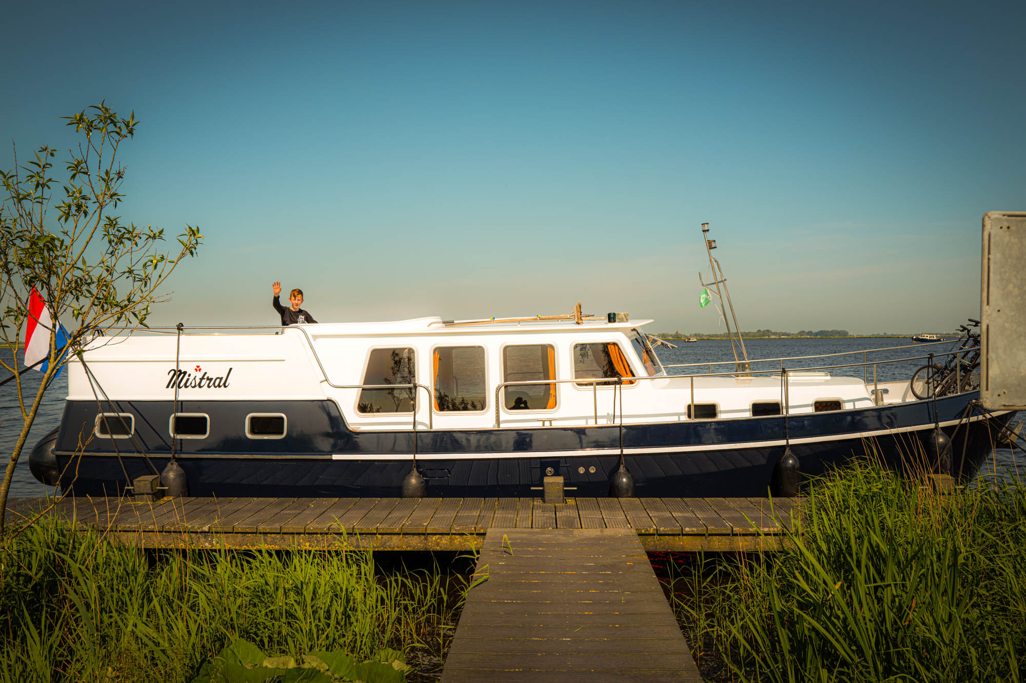A person waves from a motorboat named Mistral docked by a tranquil lake under clear blue skies.