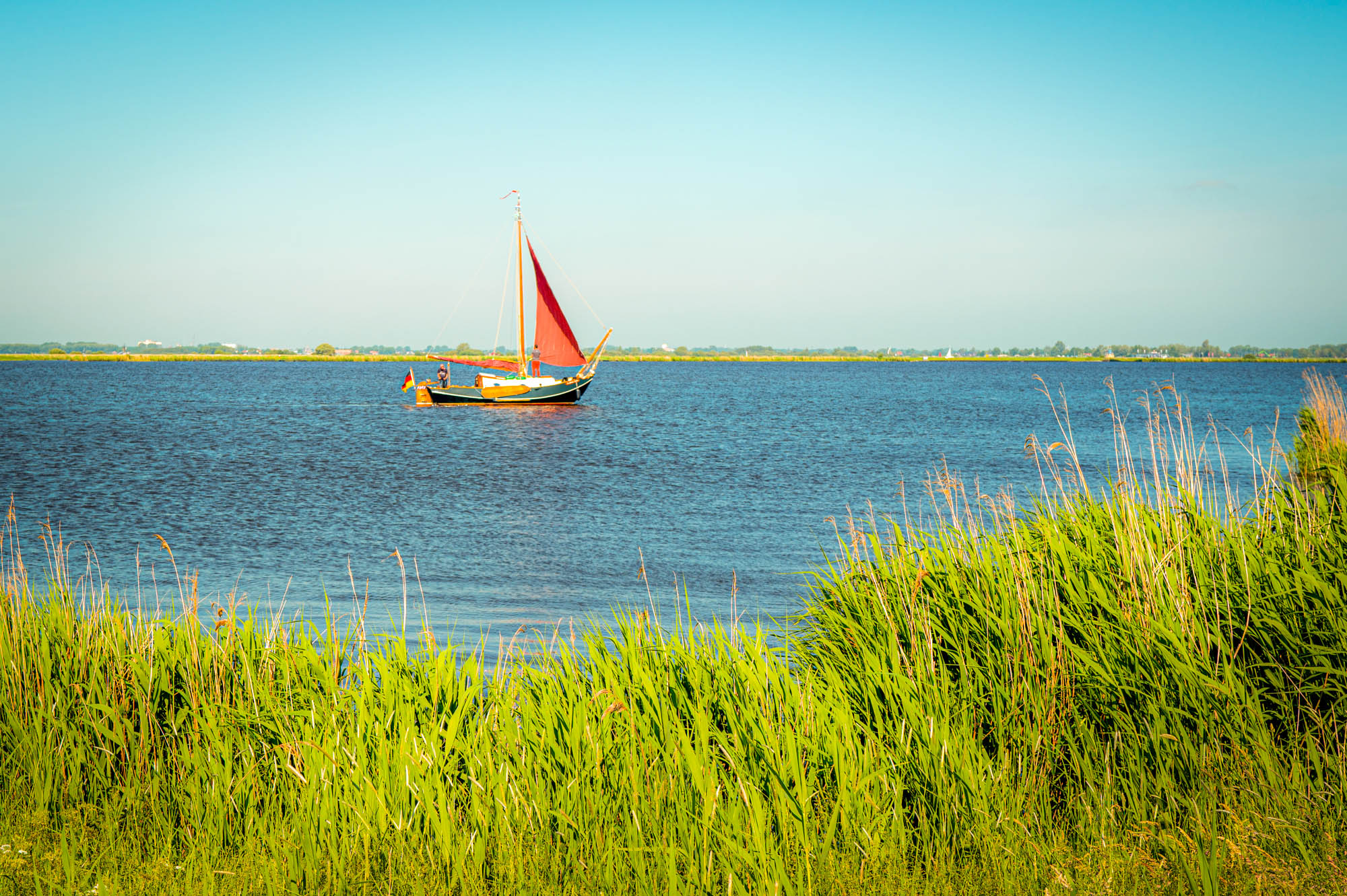 Sailboat with red sail on a tranquil lake, surrounded by green grass under a clear blue sky.