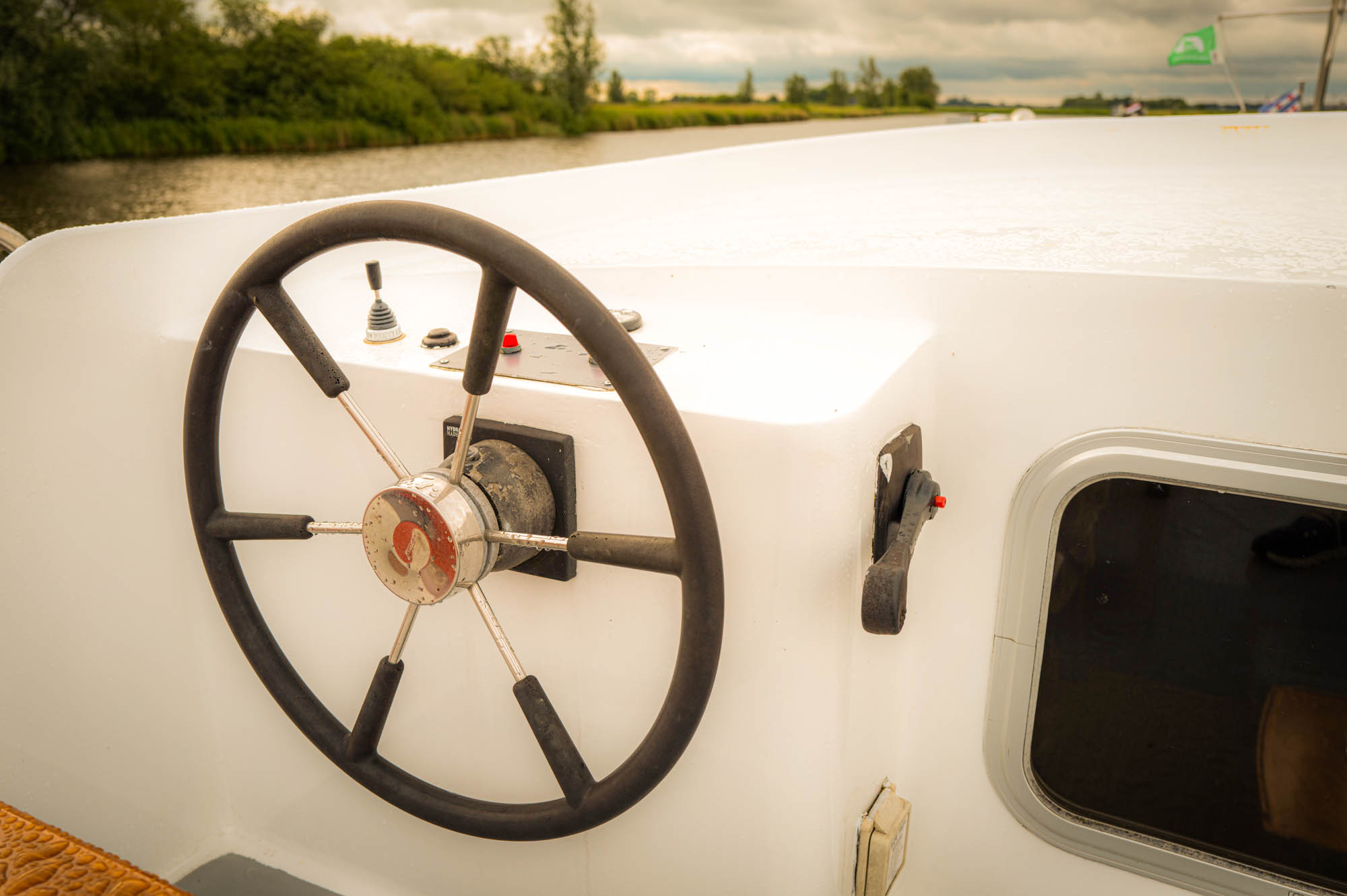 Boat steering wheel and controls on deck with river and lush green landscape in the background.