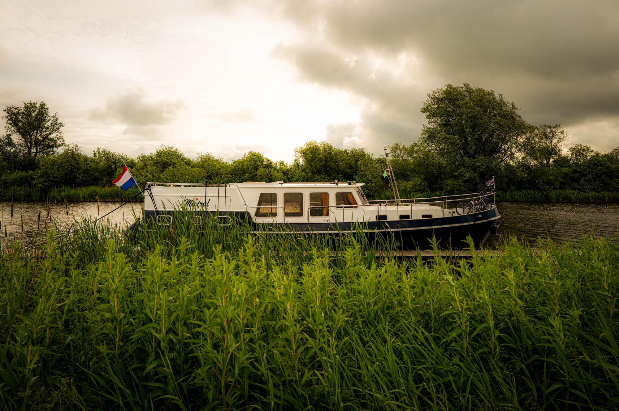 Boat docked by a river in lush green landscape under cloudy sky, with flag waving.