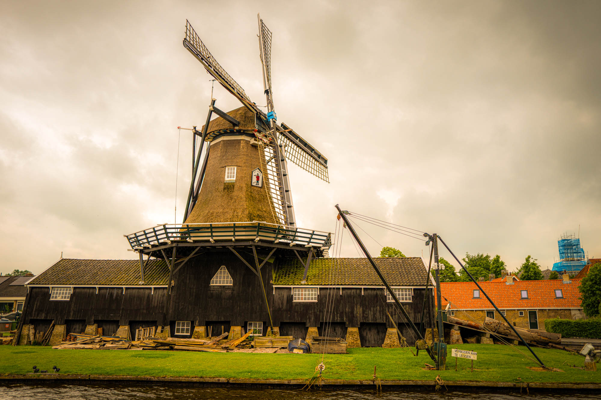 Historic Dutch windmill by riverbank under cloudy sky, surrounded by greenery and rustic buildings.