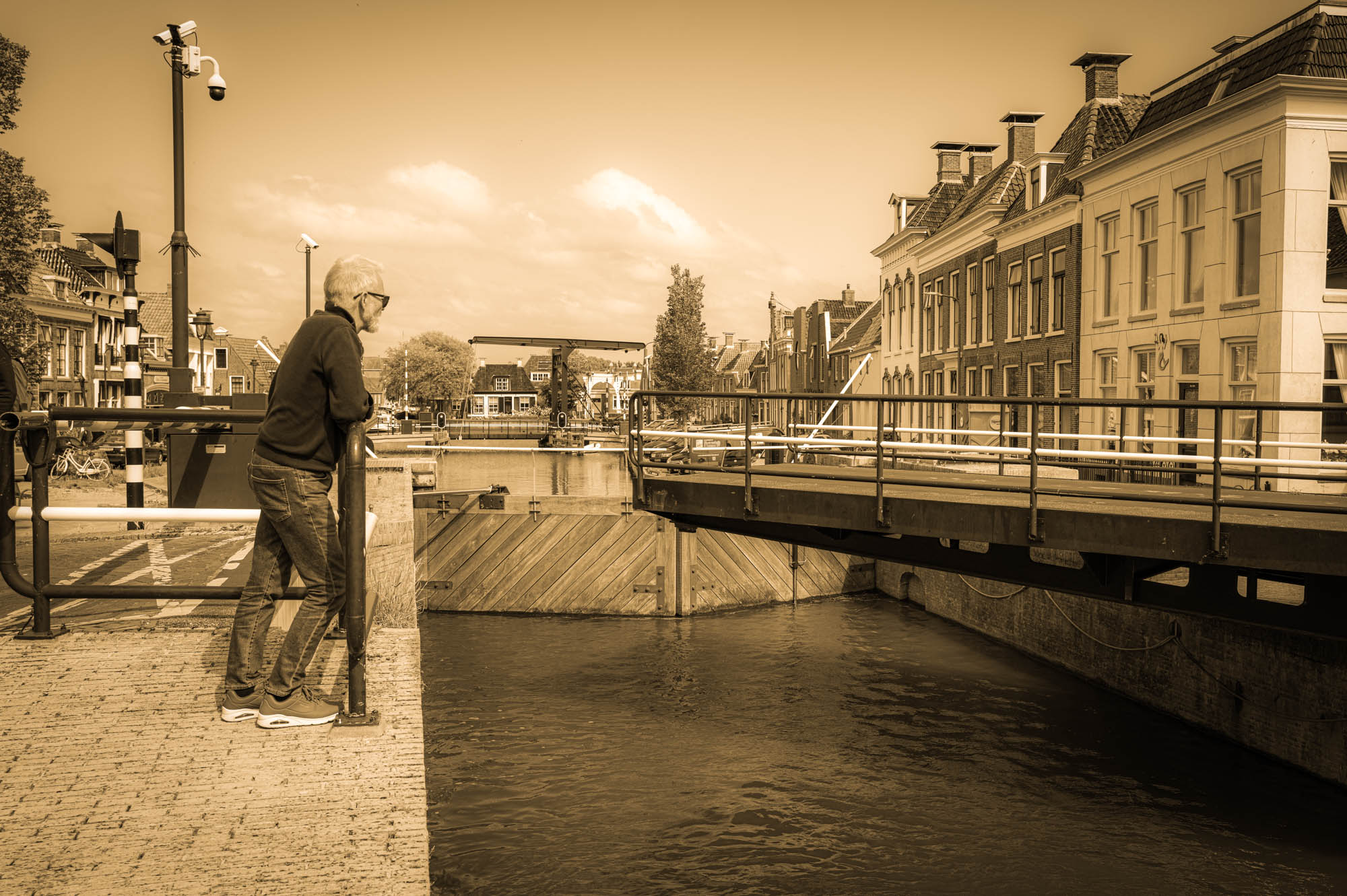 Elderly man gazes at canal near traditional houses and bridge in sepia-toned historic European town.