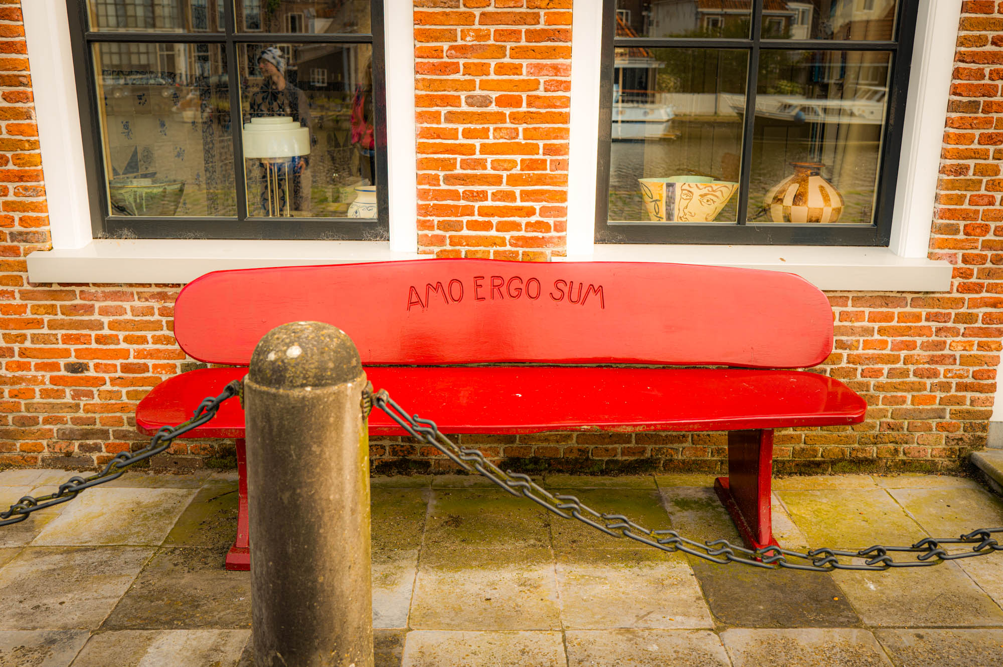 Red bench against a brick wall with Amo Ergo Sum inscribed, outside a window with vases.