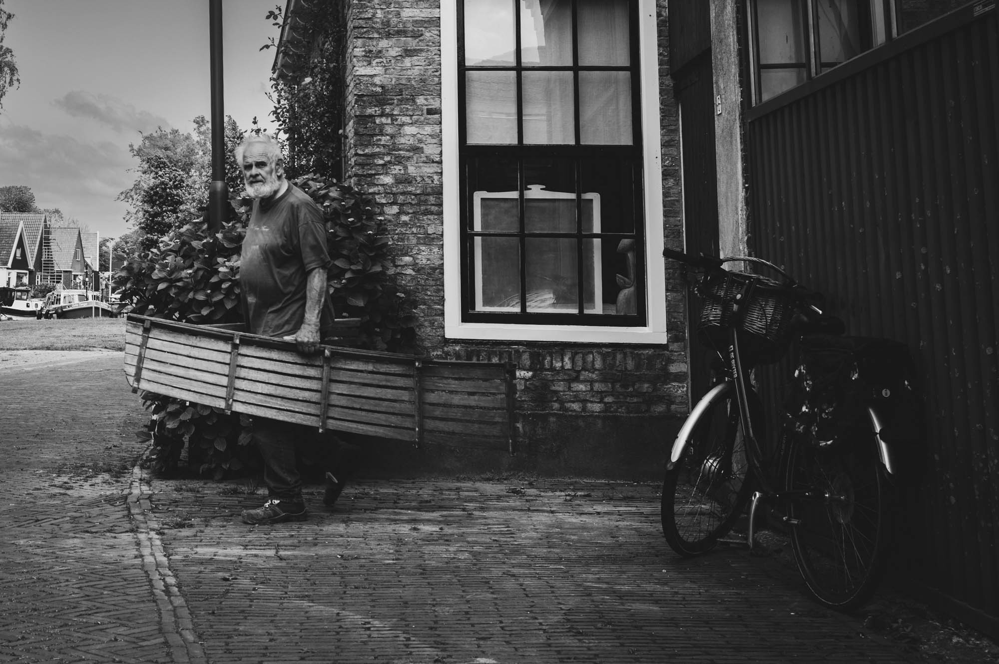 Man carrying wooden object, walking by a brick building near a parked bicycle. Black and white street scene.