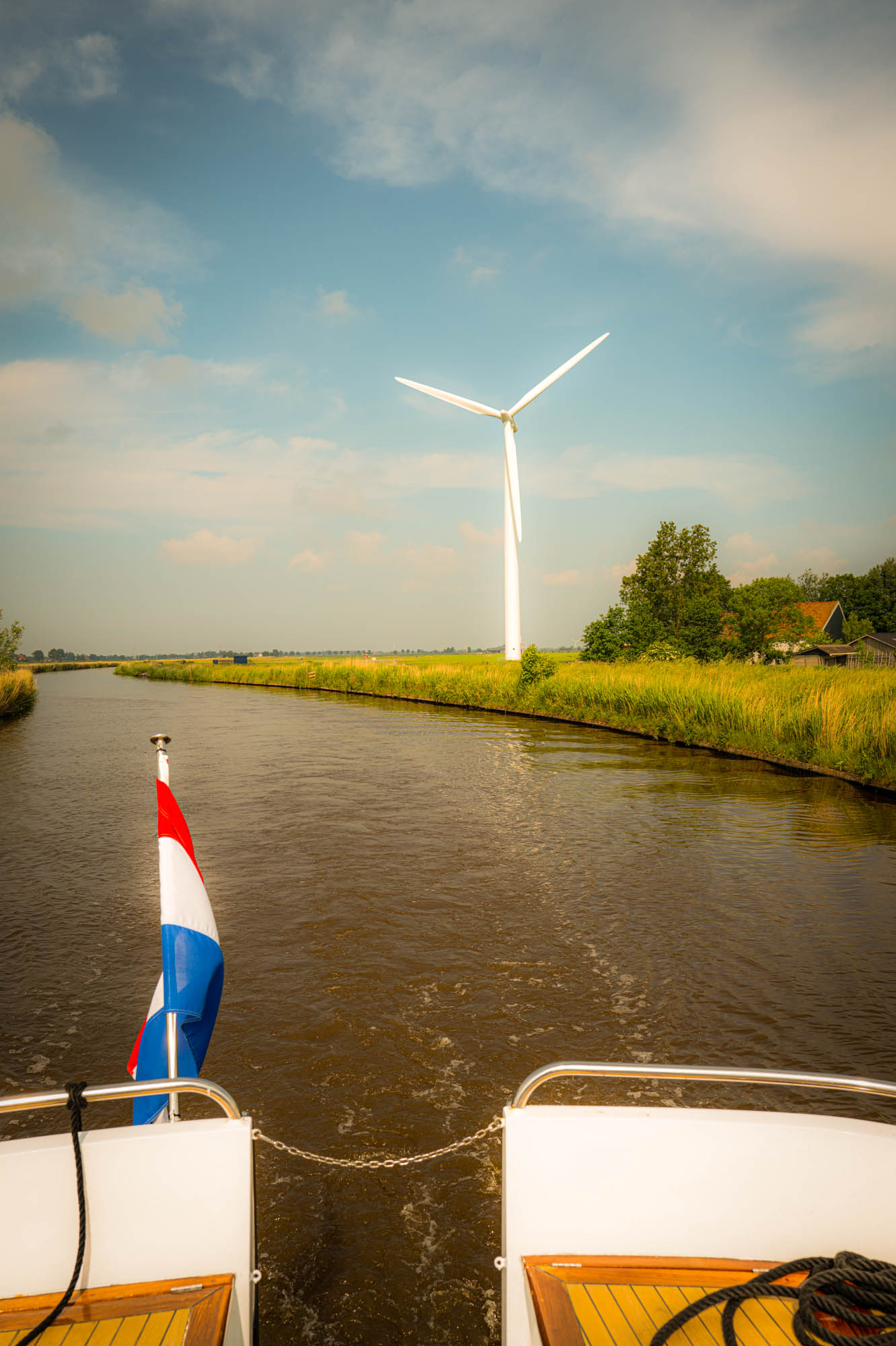 Boat with Dutch flag on a canal, wind turbine and lush greenery in the background under a clear sky.