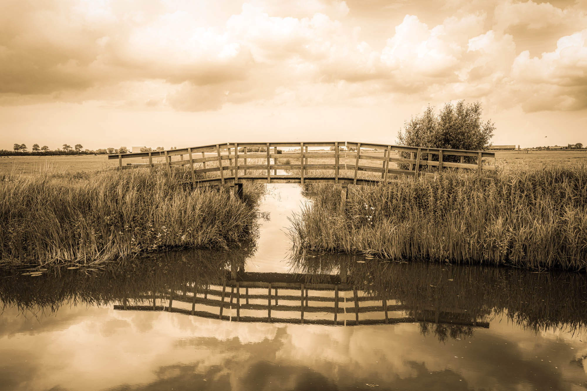 Sepia-toned landscape with wooden bridge over calm water, reflecting clouds and sky in a rural setting.