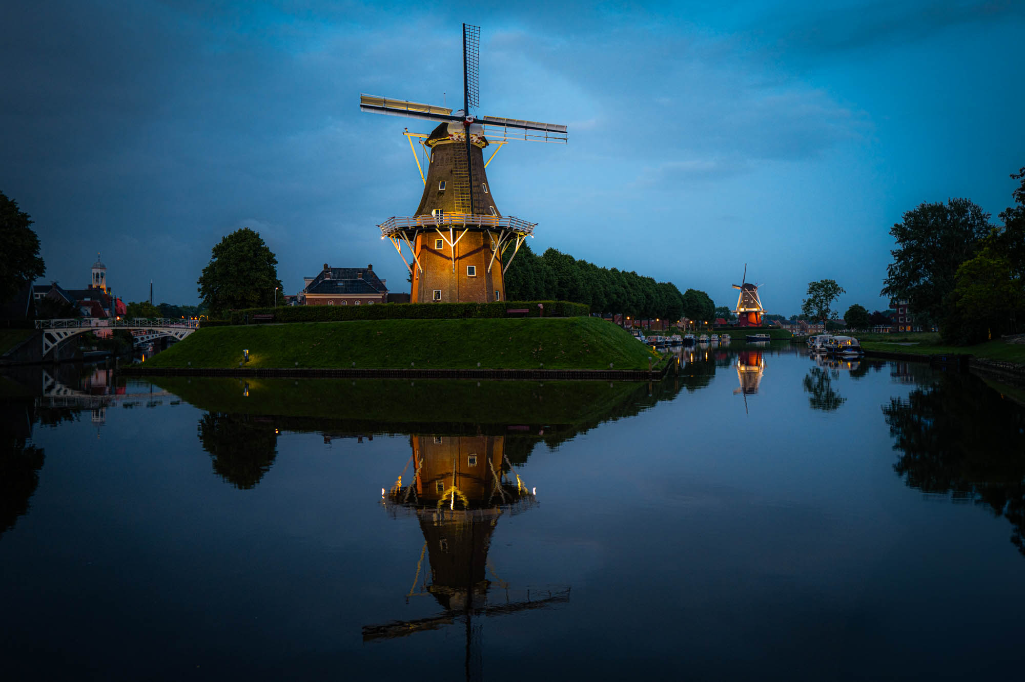 Scenic view of illuminated windmill reflecting on calm water at dusk, surrounded by trees and buildings.