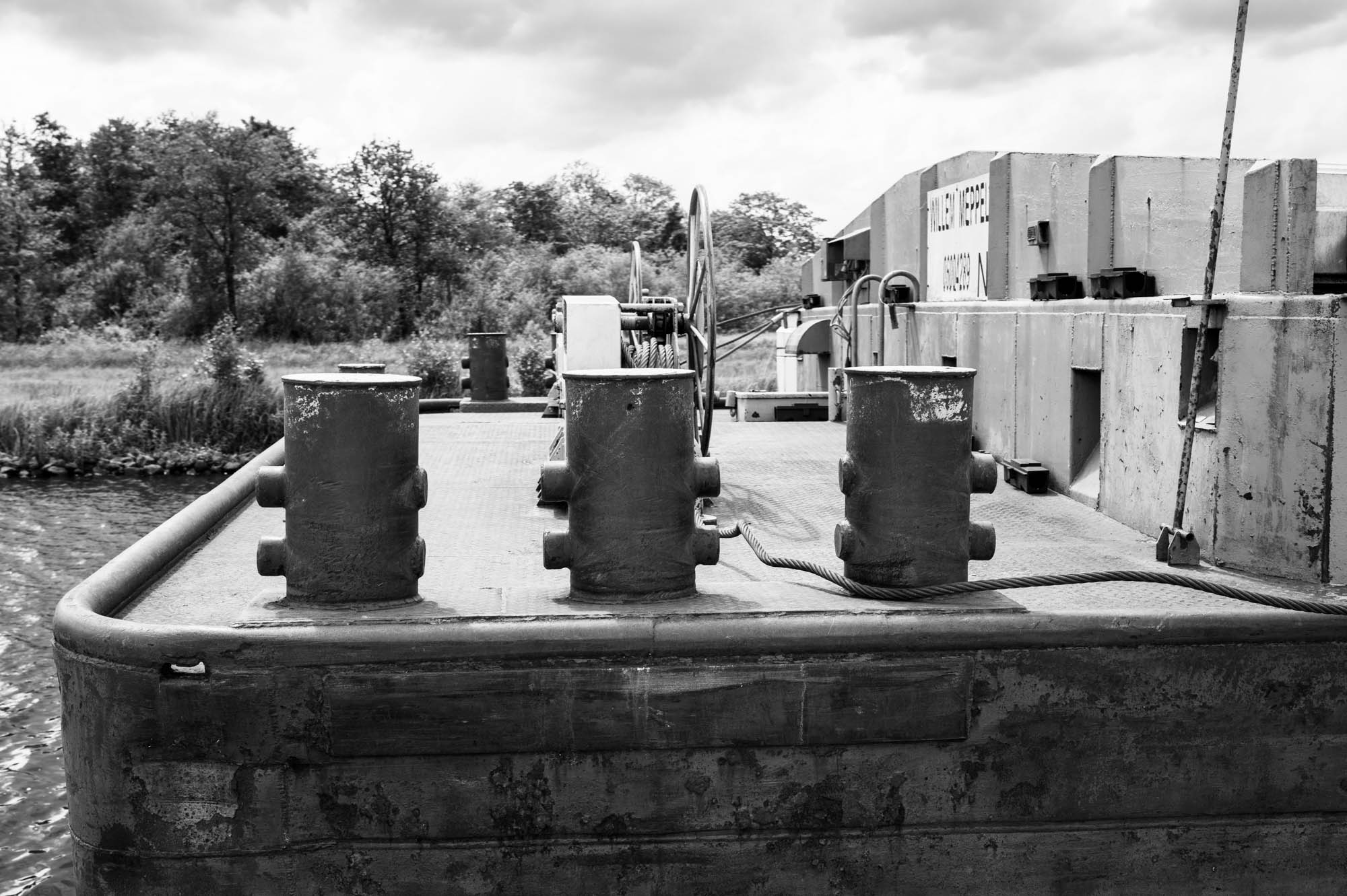 Black and white image of a barge deck with bollards and a winch near a riverside landscape.