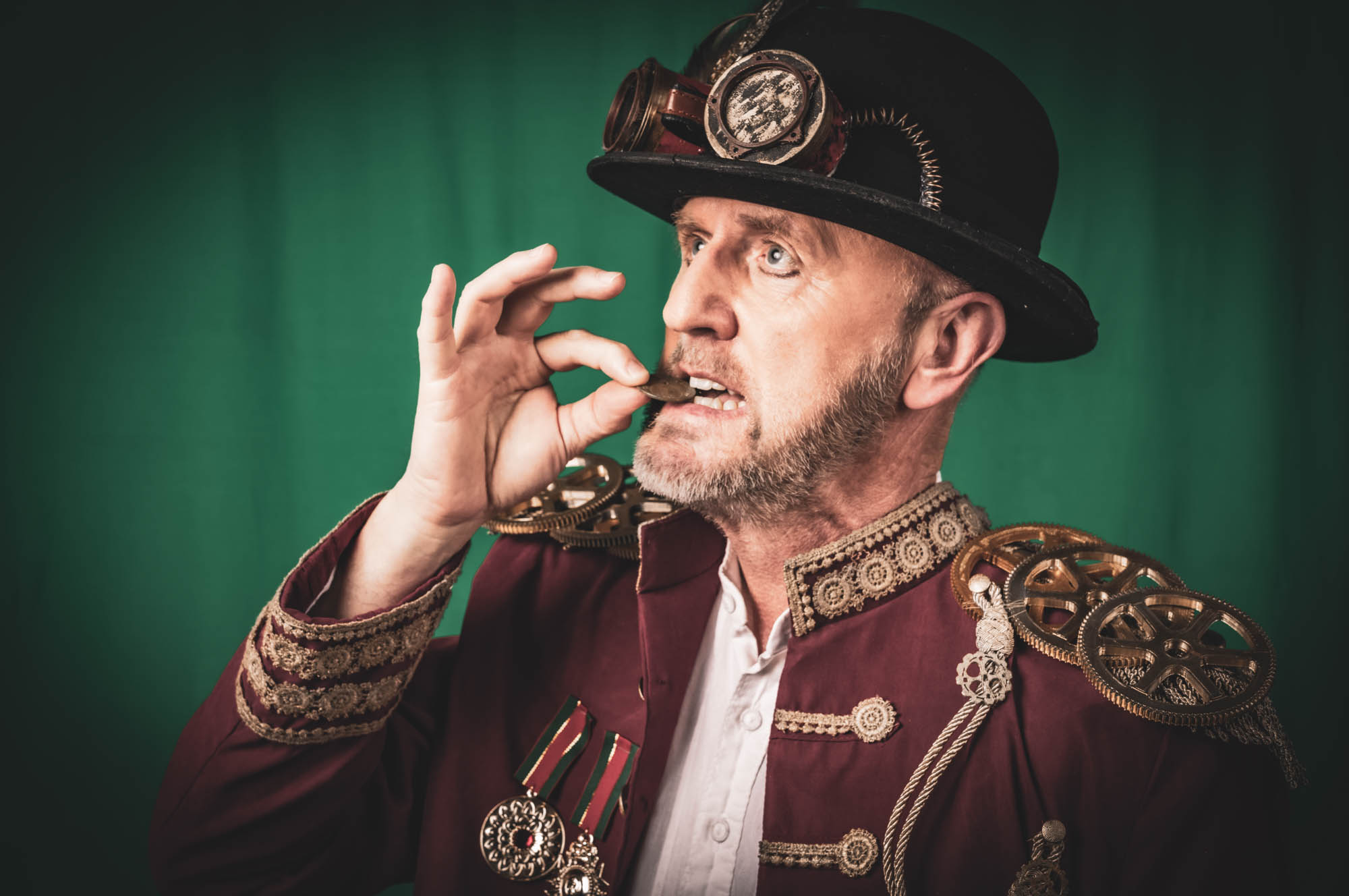 Steampunk character in vintage attire holding a coin near mouth, wearing a hat with goggles and intricate shoulder design.