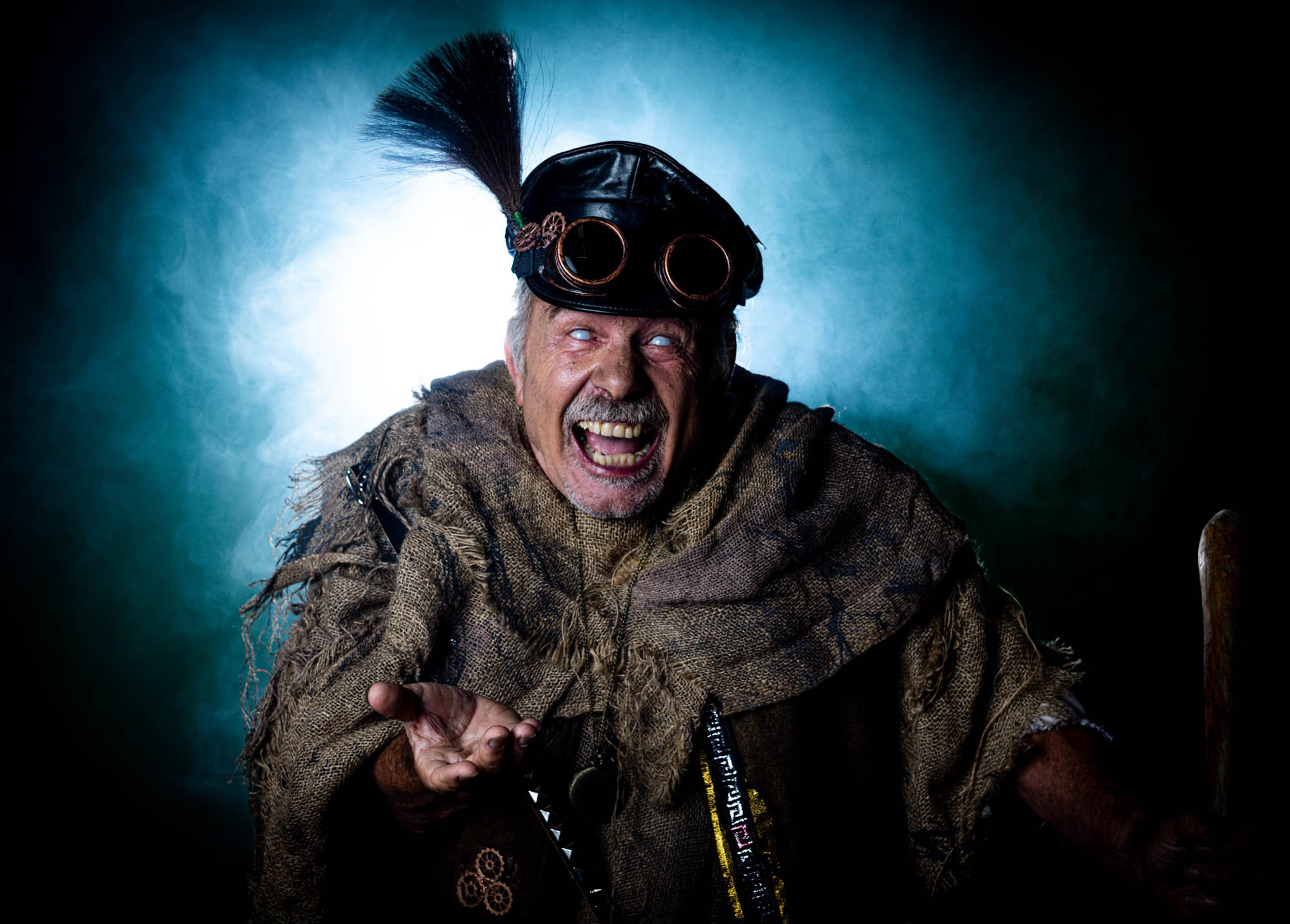 Steampunk-inspired character in rugged attire, wearing goggles and hat, making an expressive face against smoky background.