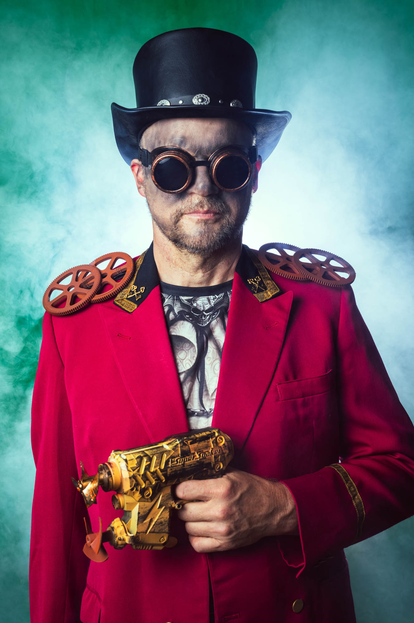 Steampunk enthusiast in red coat with goggles, hat, and golden ray gun on misty green background.
