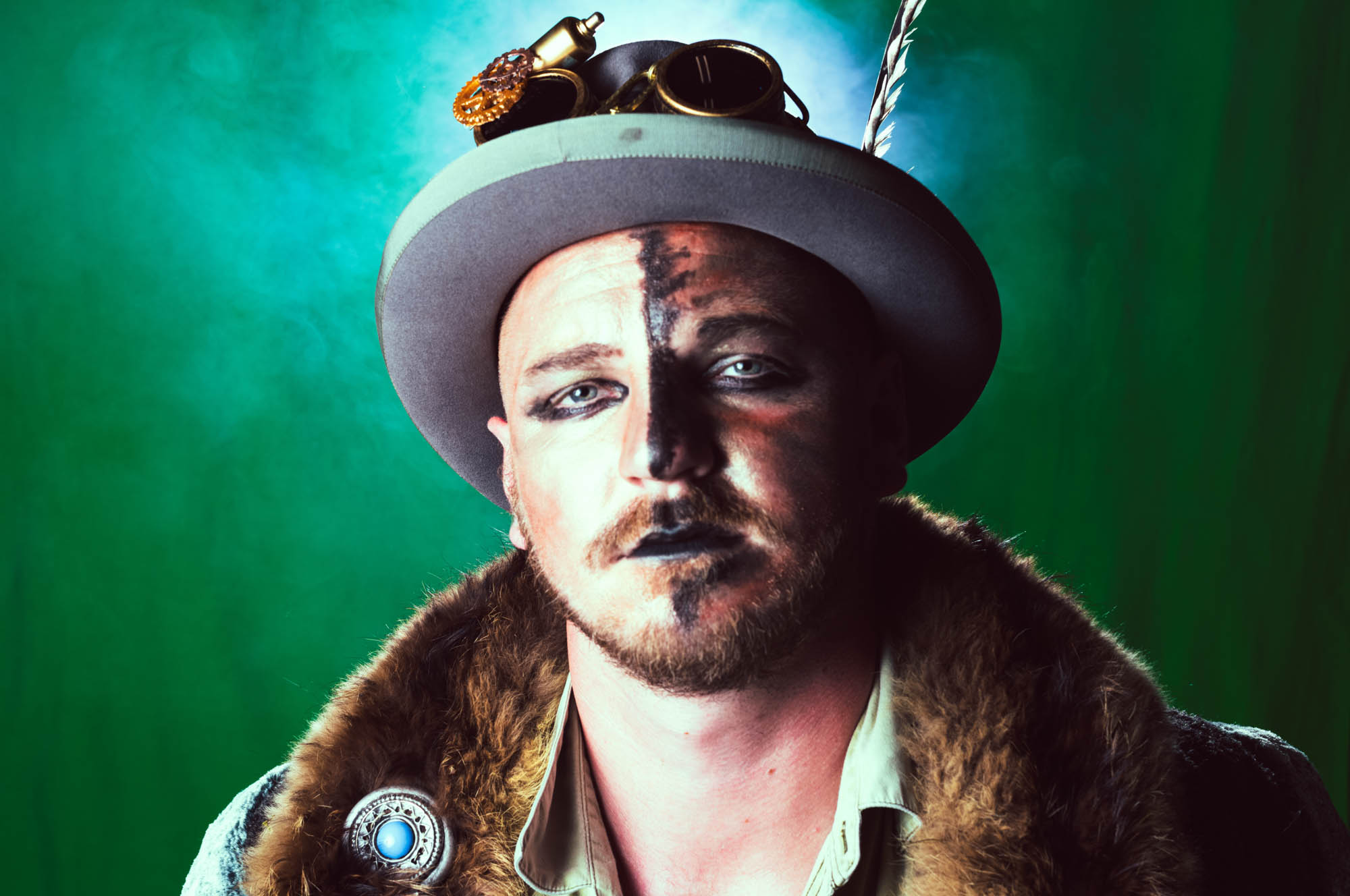 Steampunk-inspired man in a hat with feather and goggles, wearing fur coat, against a green background.