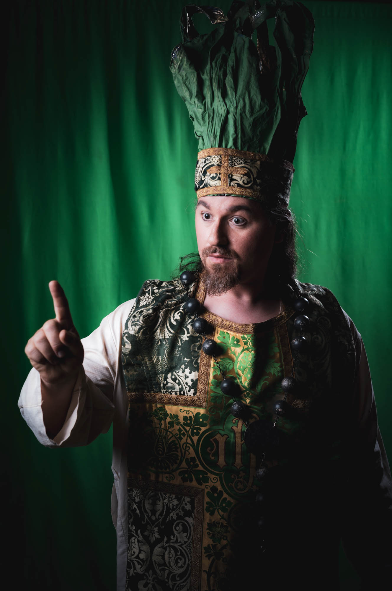 Man in ornate medieval costume and tall hat gesturing, against a green background.