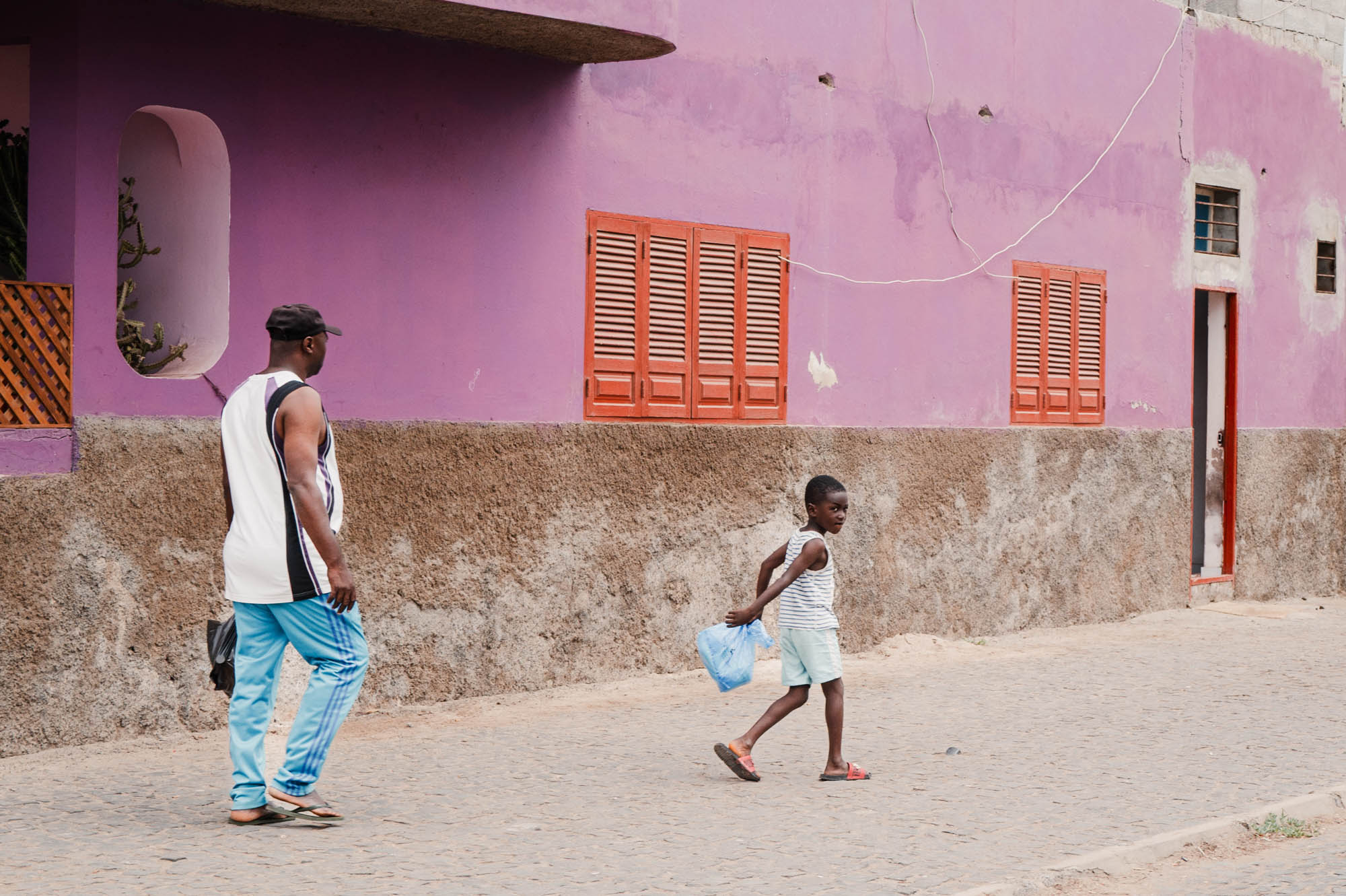 Man and child walking by a colorful house with a purple wall and orange shutters, child holds a blue bag.