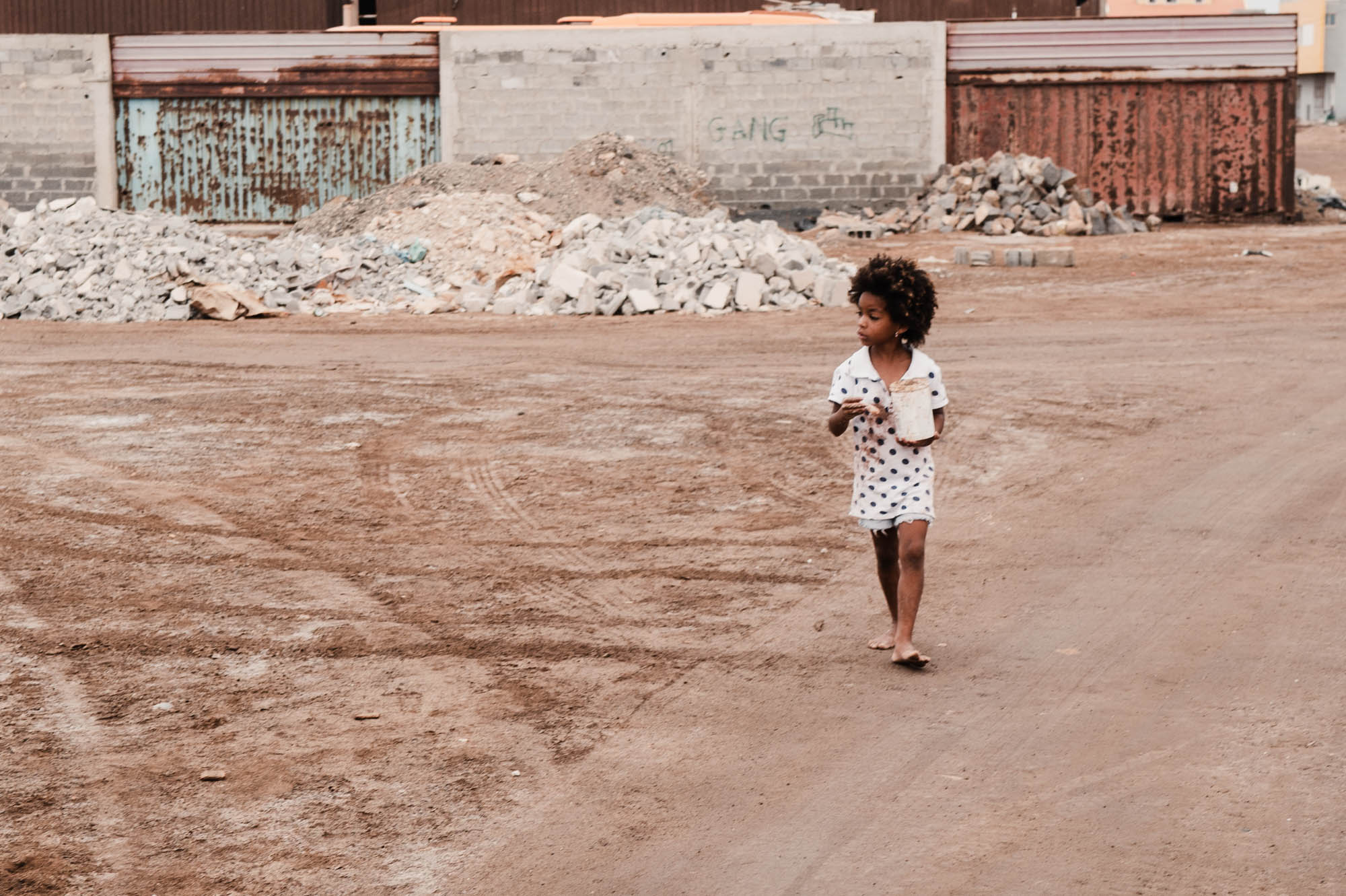 Child walking barefoot on a dirt path near rubble and corrugated steel, holding a container, in an urban setting.