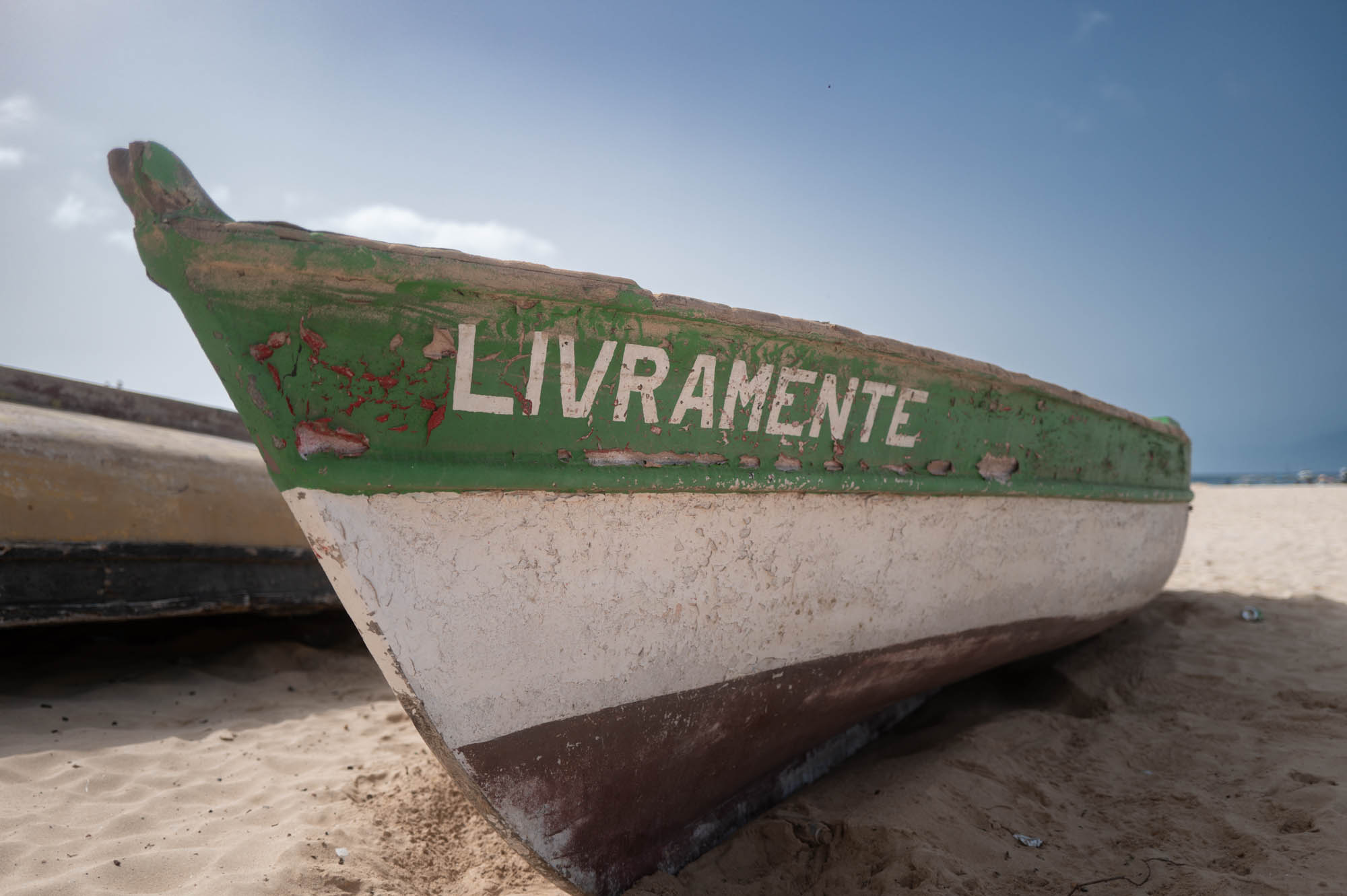 Old green boat named LIVRAMENTE resting on sandy beach under clear blue sky.