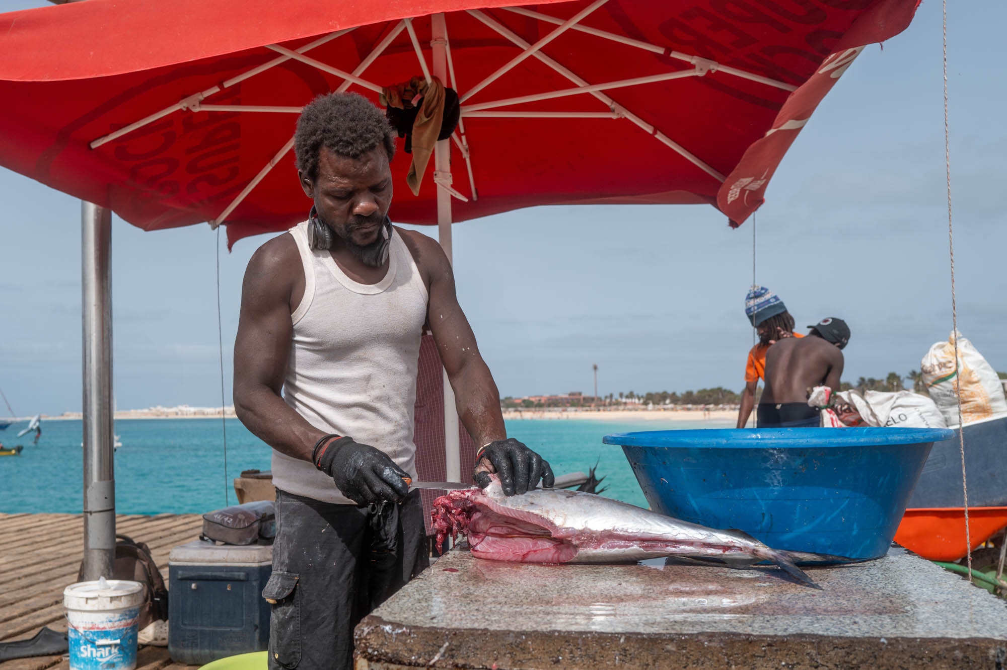 Man filleting fish under red umbrella by the ocean, beach and boats in the background.