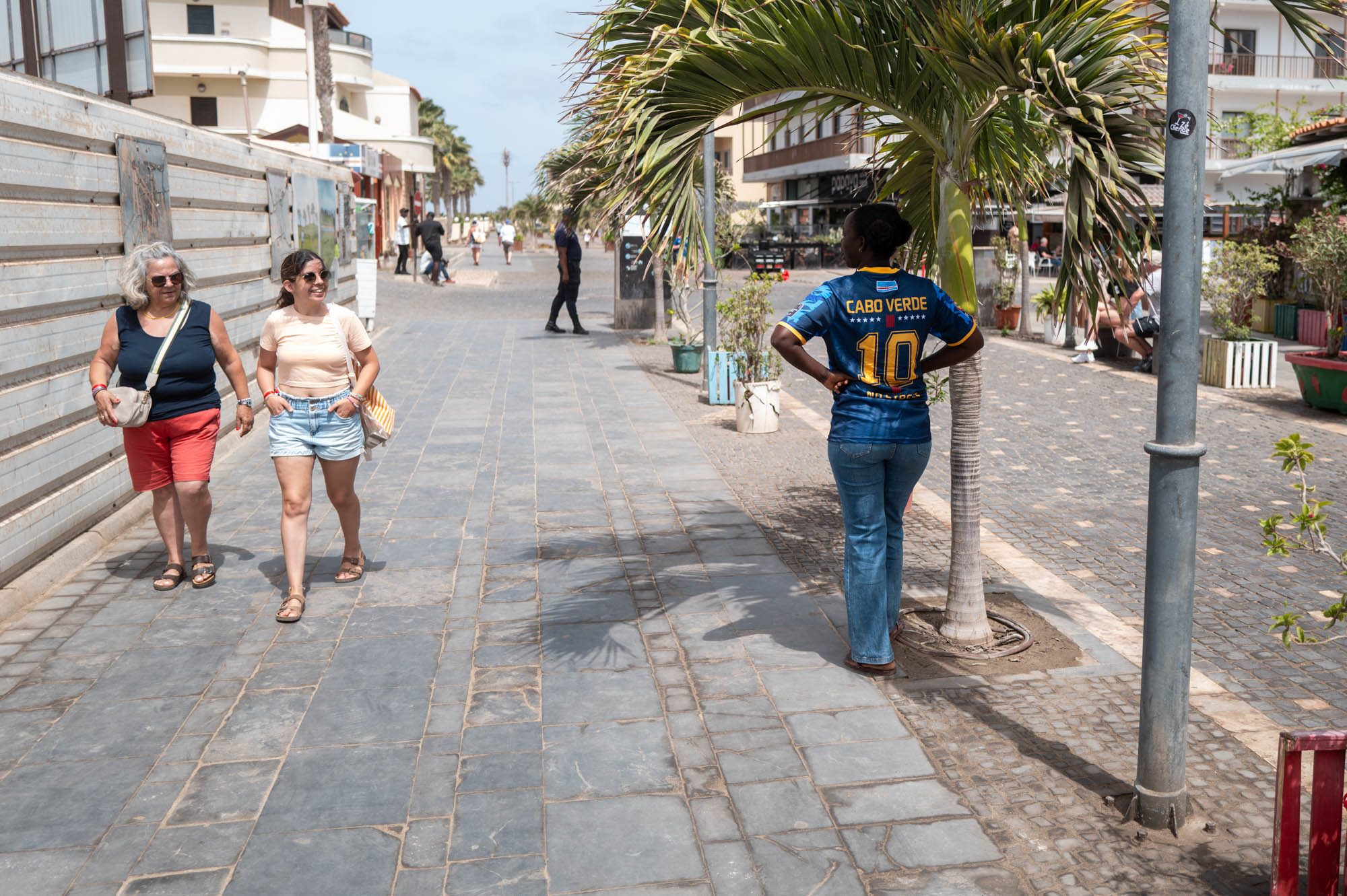 People walking on a sunny street near a palm tree, one wearing a Cabo Verde shirt, showcasing urban life.
