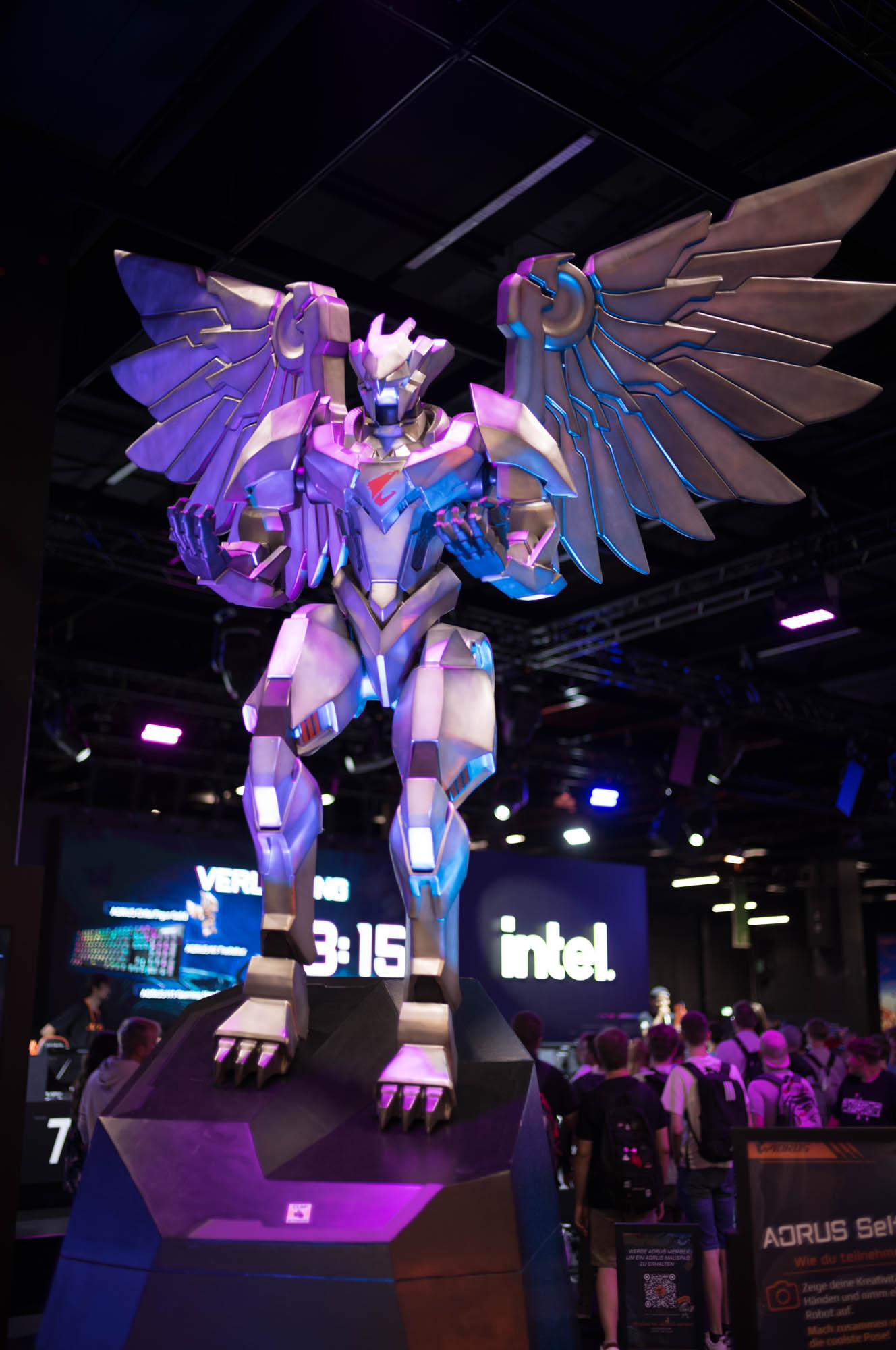 Statue of futuristic winged robot on display at a tech event with colorful lighting and crowd in the background.