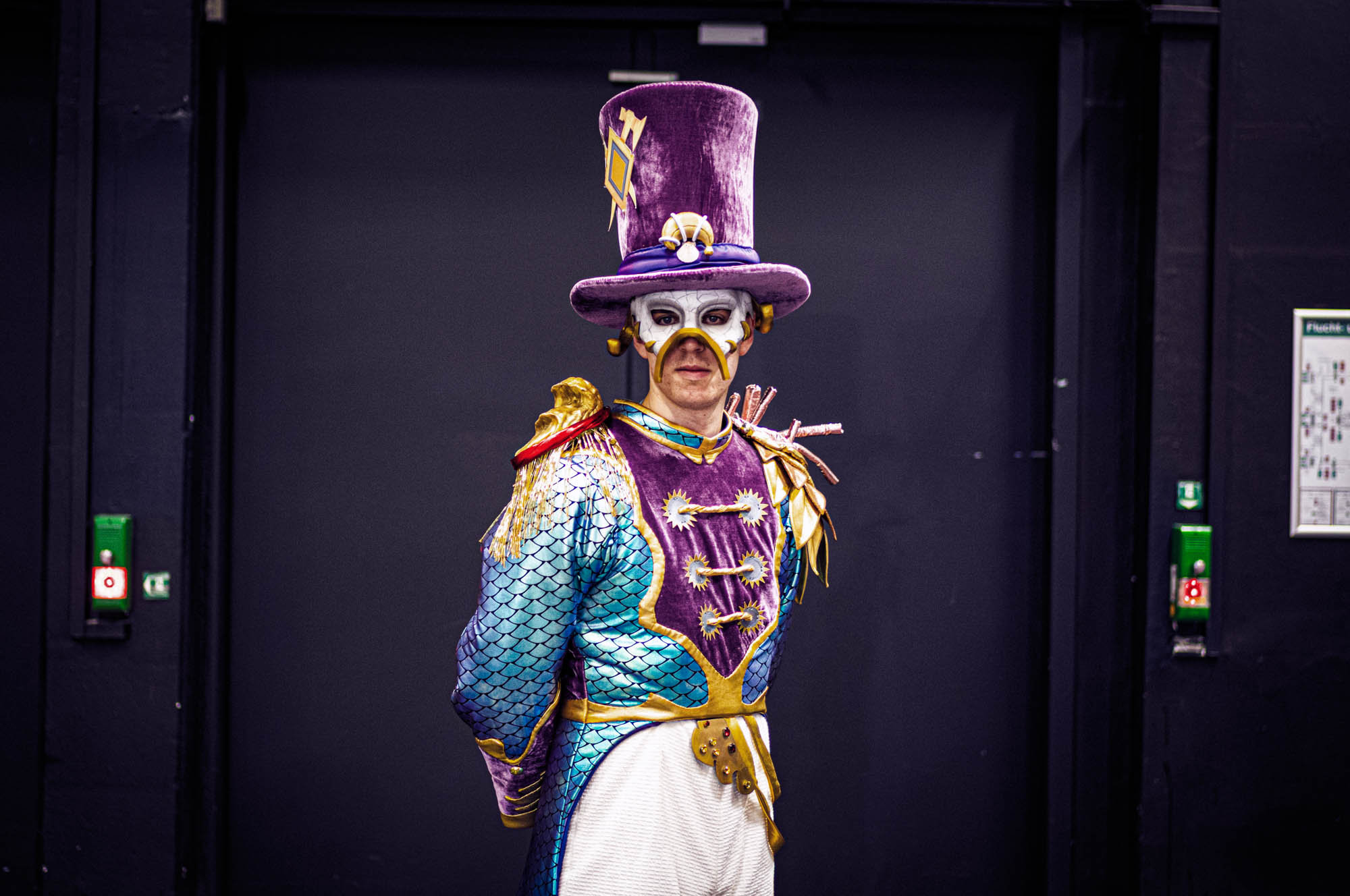 Person in colorful, theatrical costume with a purple top hat and mask in front of a dark background.